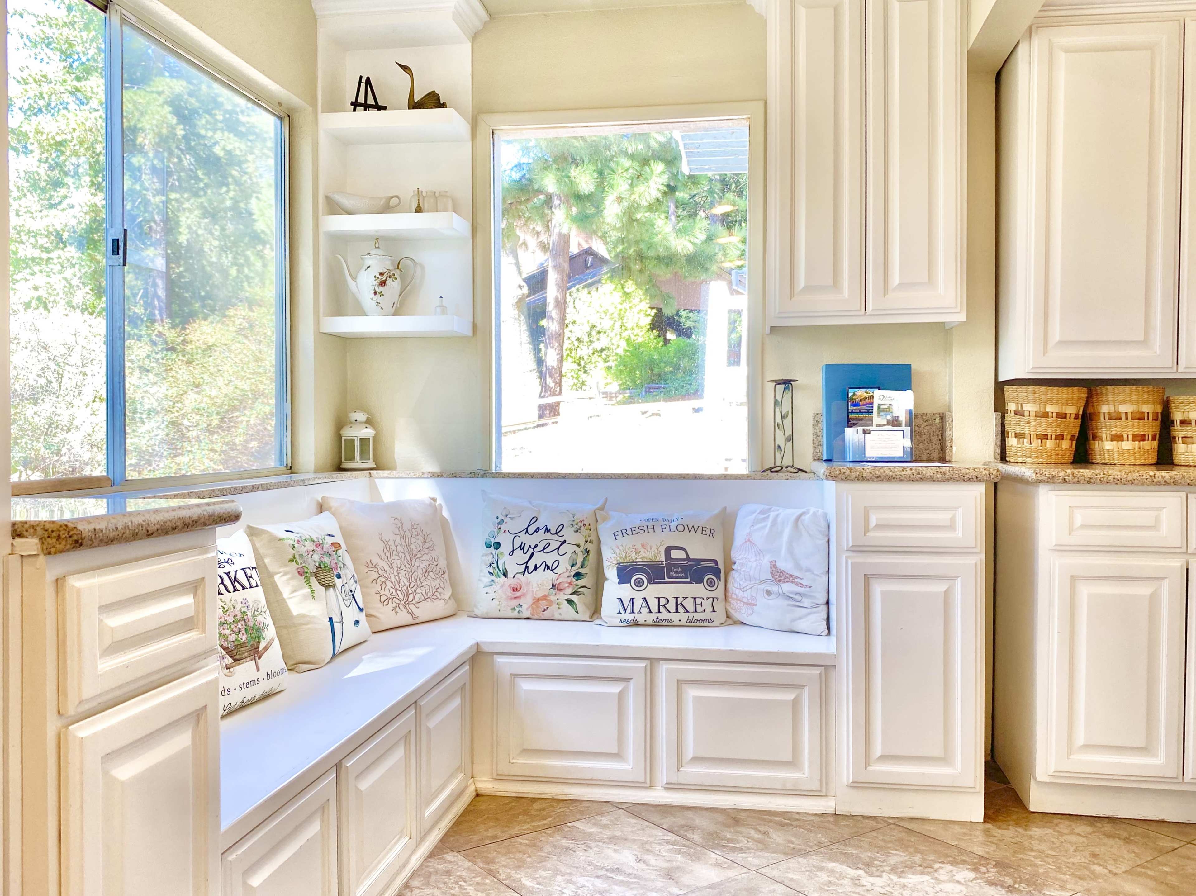 The image shows a sunny kitchen corner with a built-in bench featuring decorative pillows and white cabinetry alongside a large window.