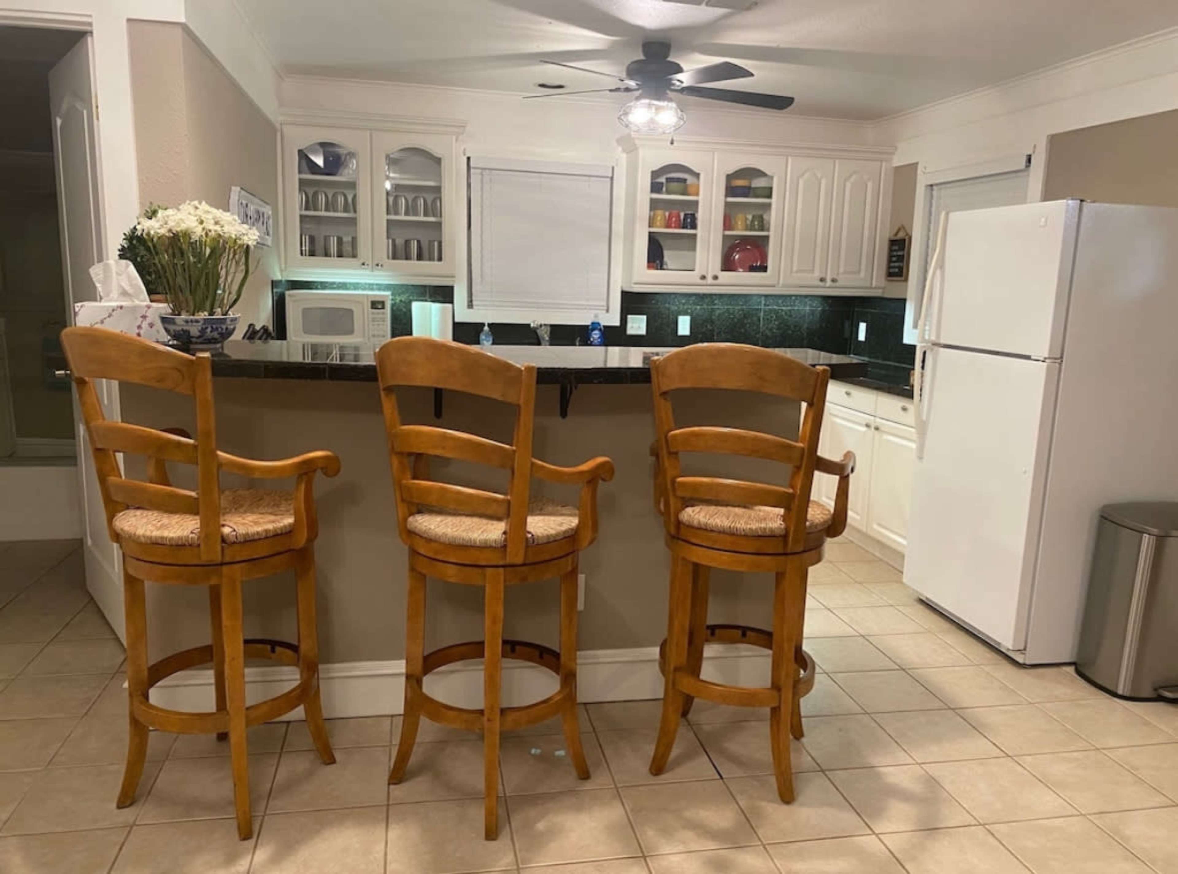A kitchen with three wooden bar stools lined up at a counter, alongside white cabinetry, a refrigerator, and a microwave.