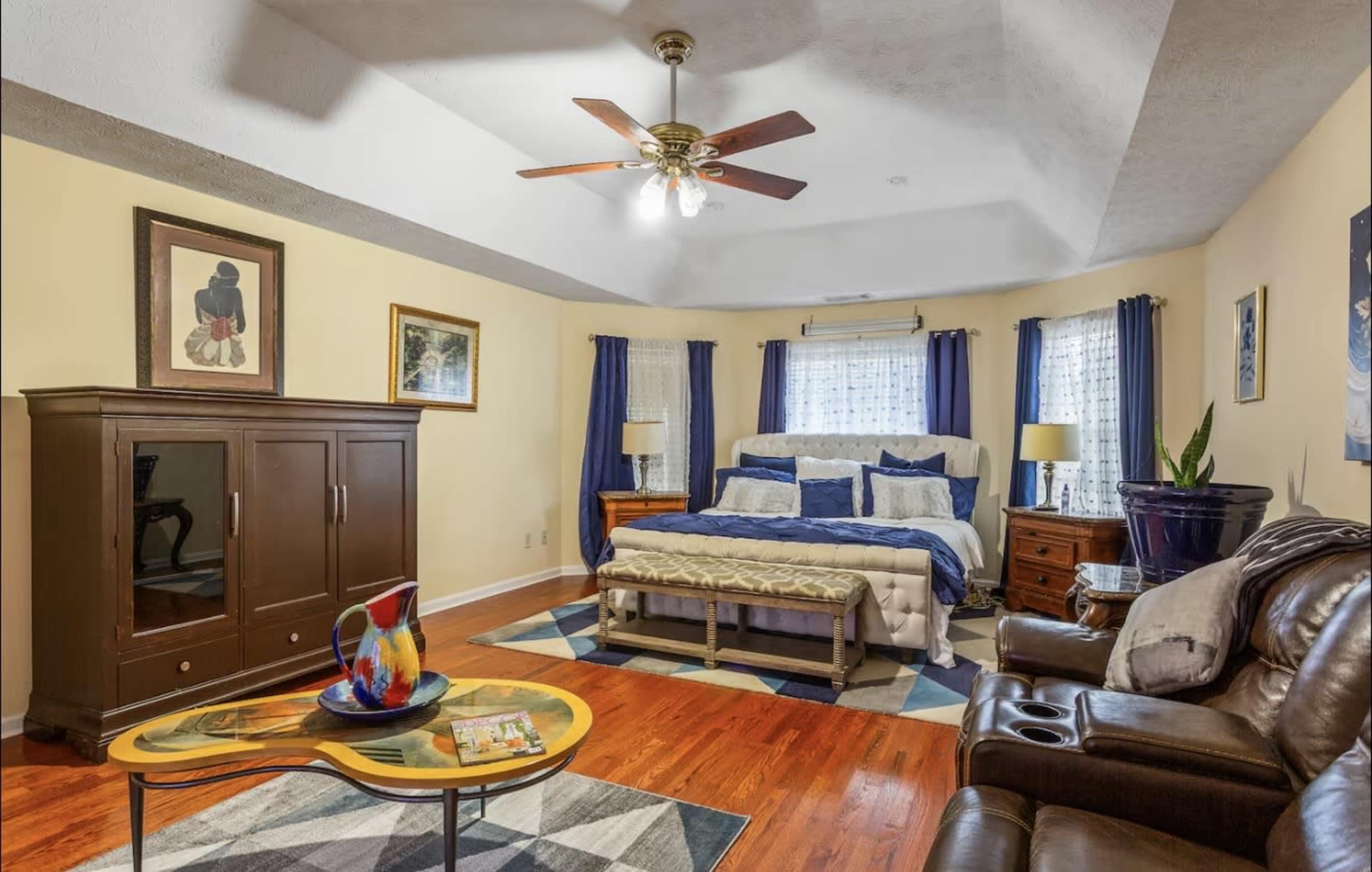 The image shows a spacious bedroom with a large bed, wooden furniture, and a seating area featuring a recliner and a coffee table.