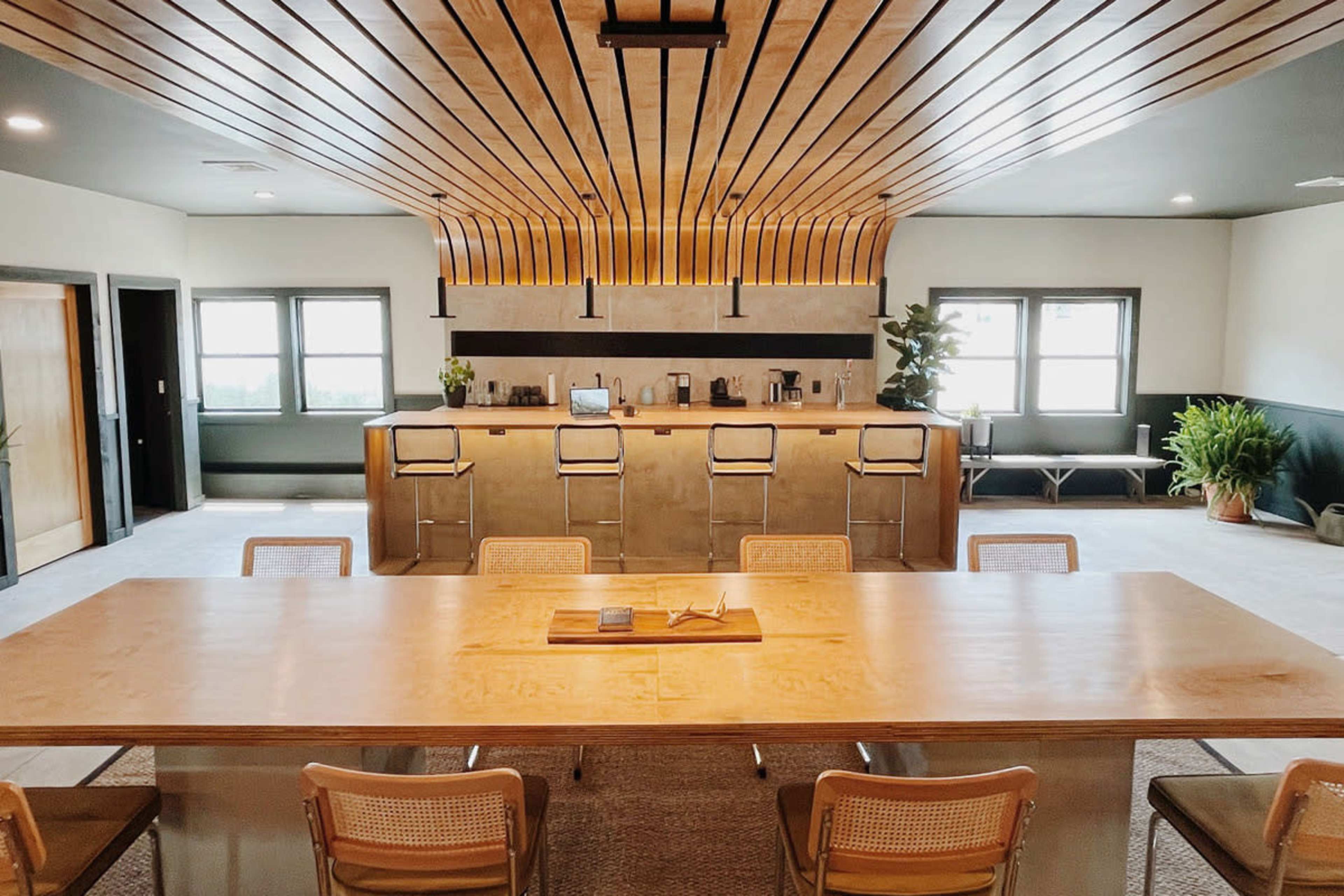 A modern office breakroom featuring a long wooden table, several chairs, and a kitchen area with bar stools, cabinets, and coffee machines.