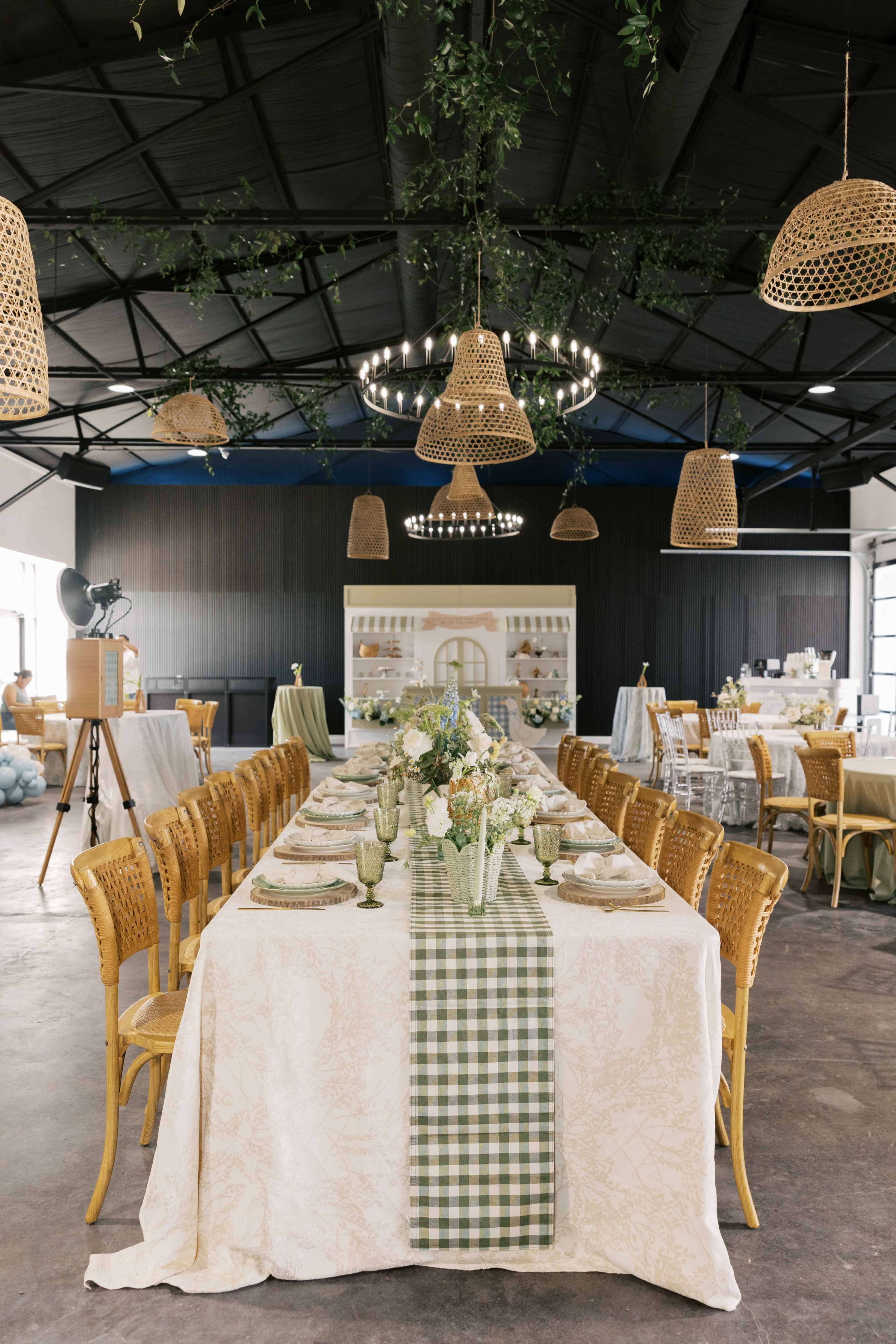 A long dining table is set with decorative tableware and floral arrangements under a ceiling adorned with woven pendants and greenery.