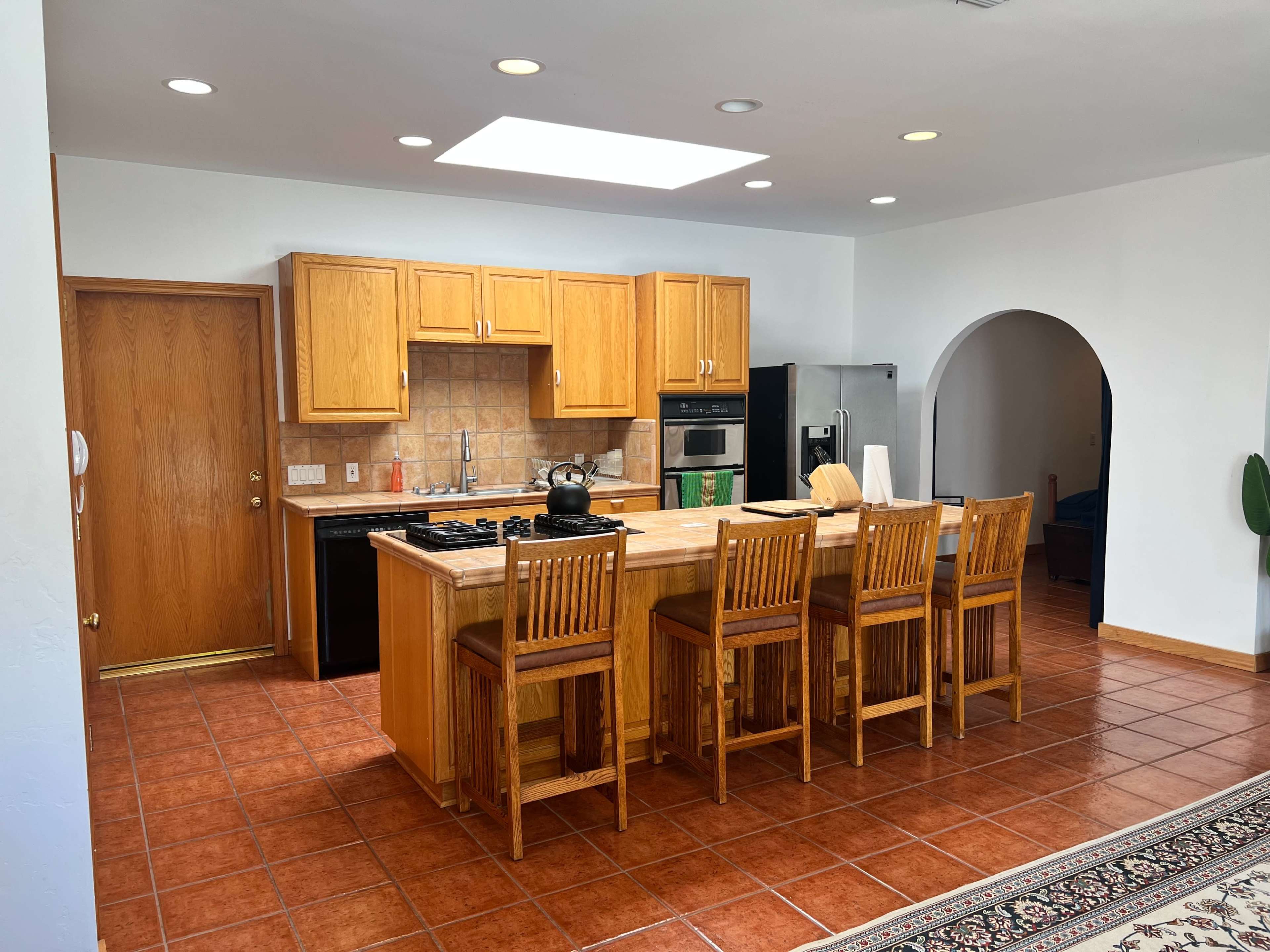 The image shows a spacious kitchen with wooden cabinetry, a central island with seating for four, and modern appliances.