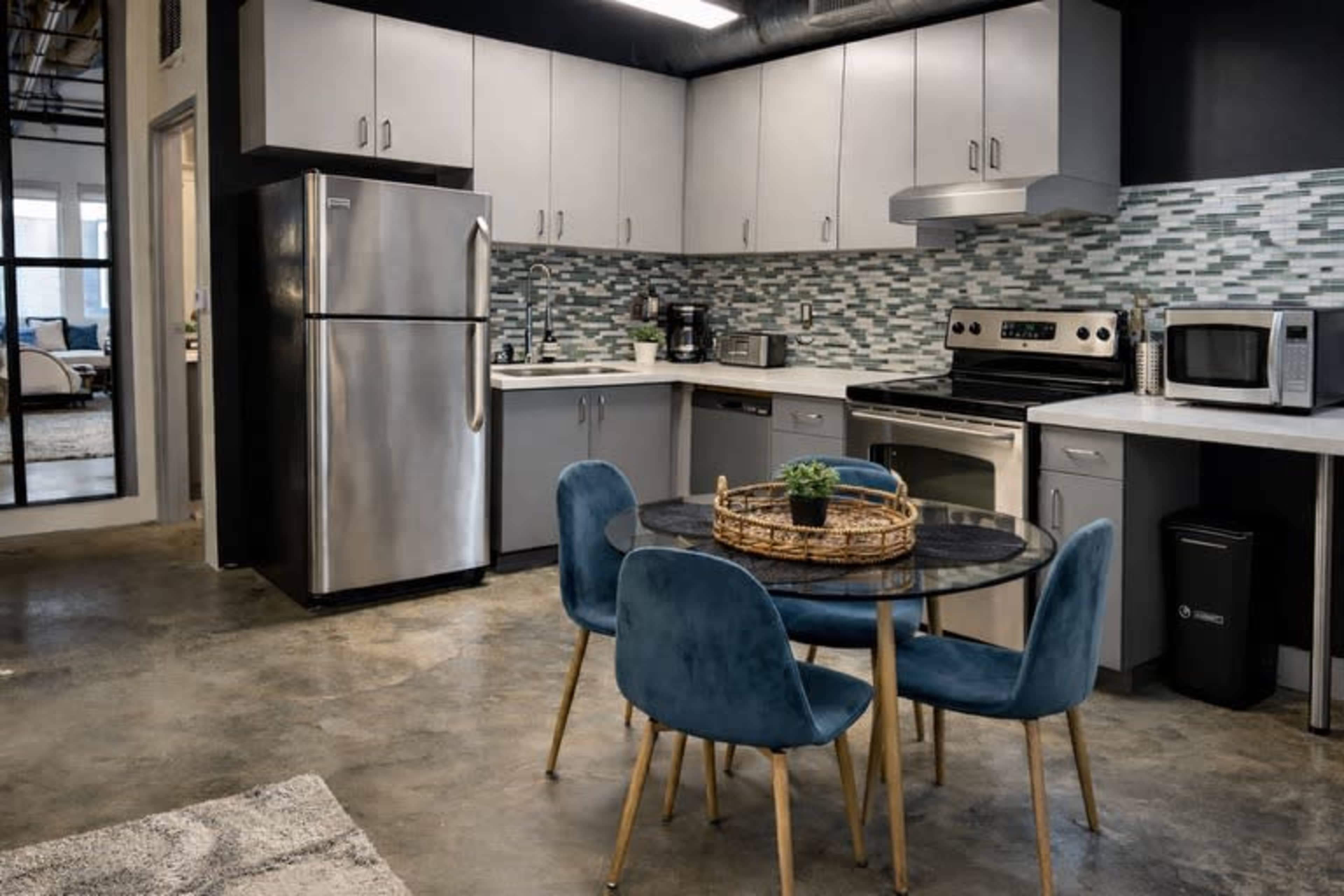 The image shows a modern kitchen with stainless steel appliances, gray cabinetry, and a round dining table surrounded by four blue chairs.