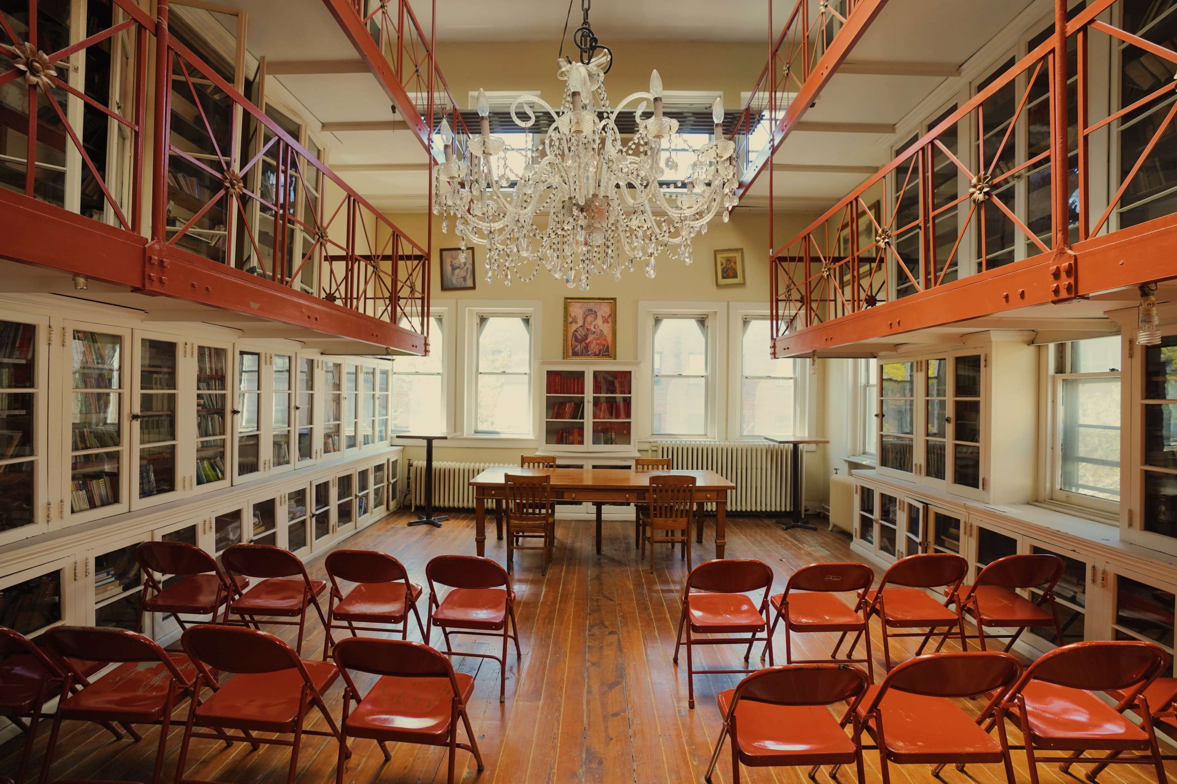 A spacious library featuring two levels of bookshelves, a chandelier overhead, and rows of red chairs arranged in front of a central table.