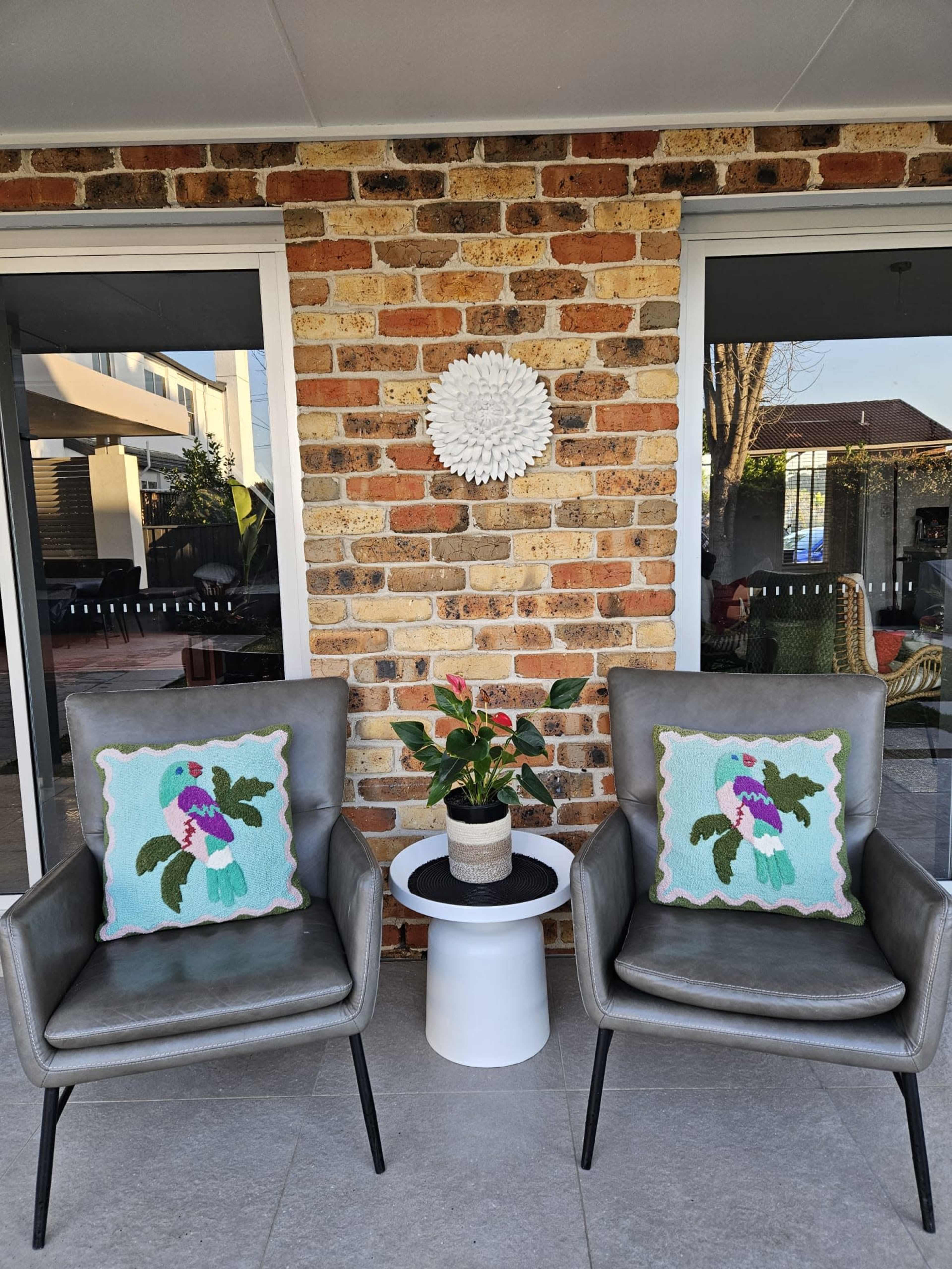 Two gray chairs with decorative cushions face each other beside a small white table, set against a brick wall with large windows.
