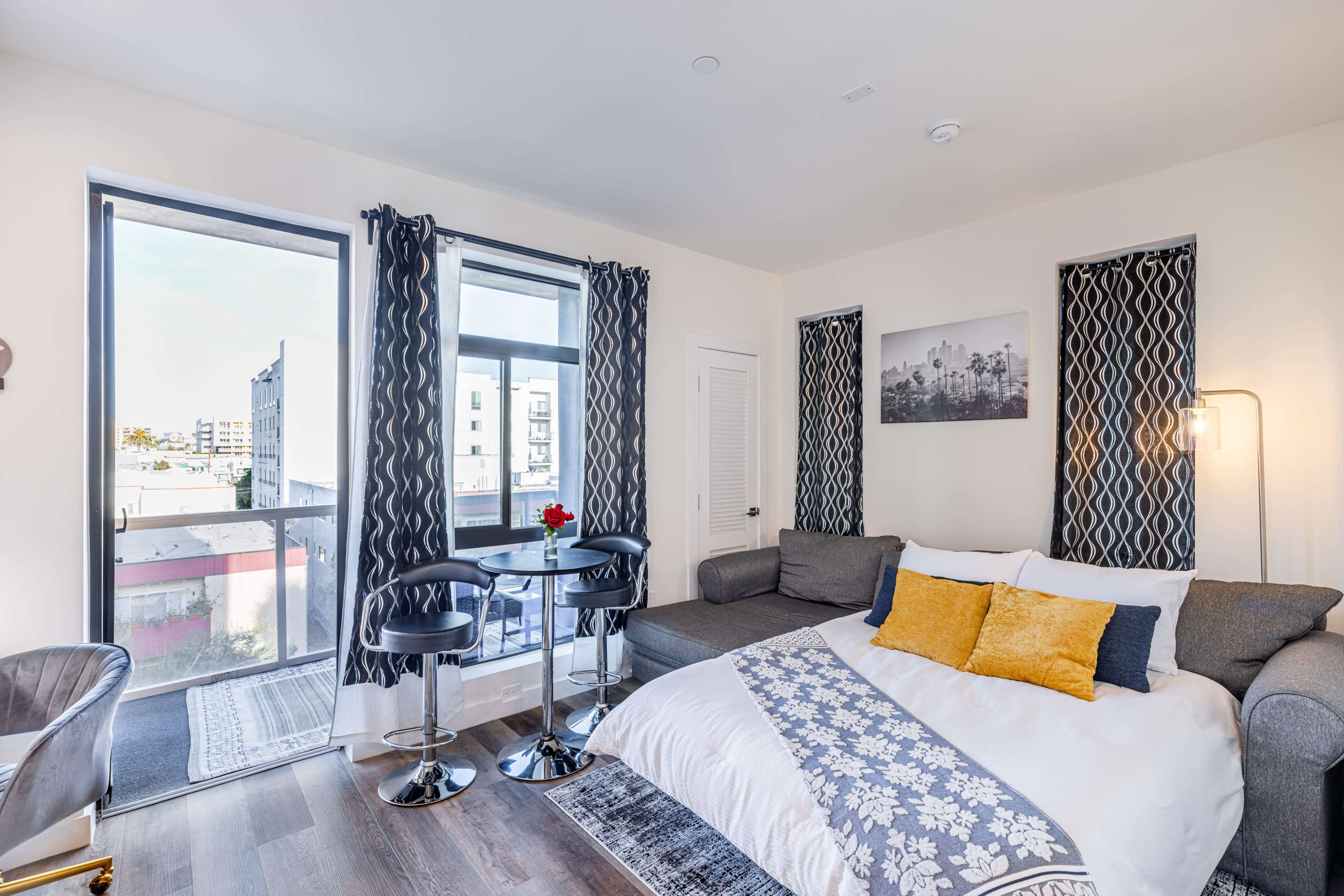 A well-lit studio apartment features a bed, a small dining table with two chairs, and large windows with views of the outside.