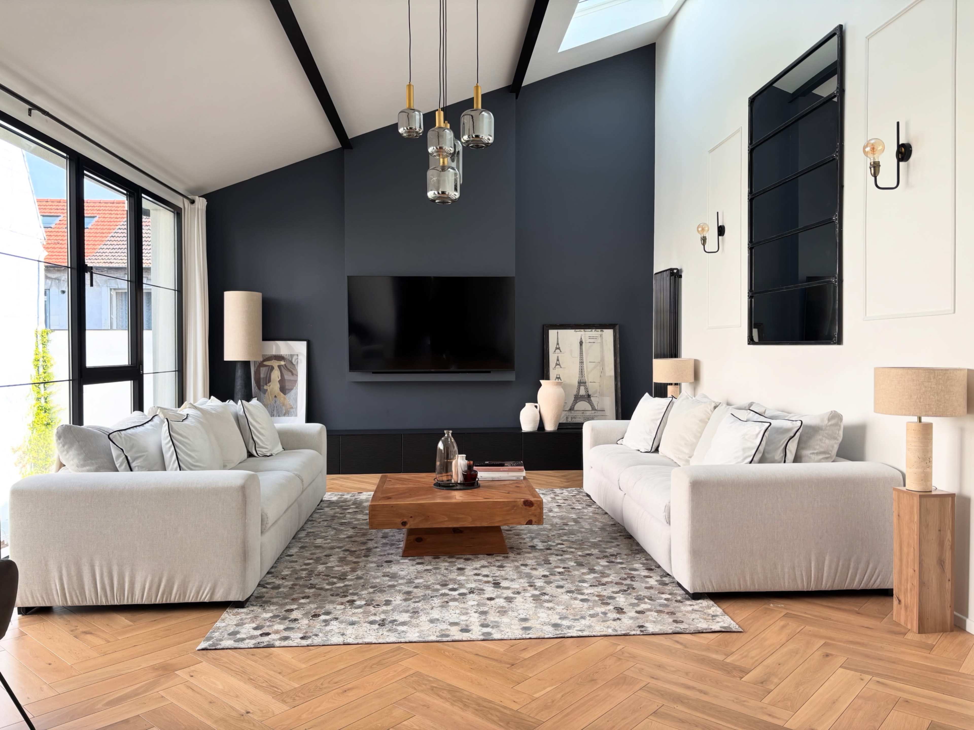 The living room features two light-colored sofas, a wooden coffee table, and a large television mounted on a dark accent wall, with large windows allowing natural light to fill the space.