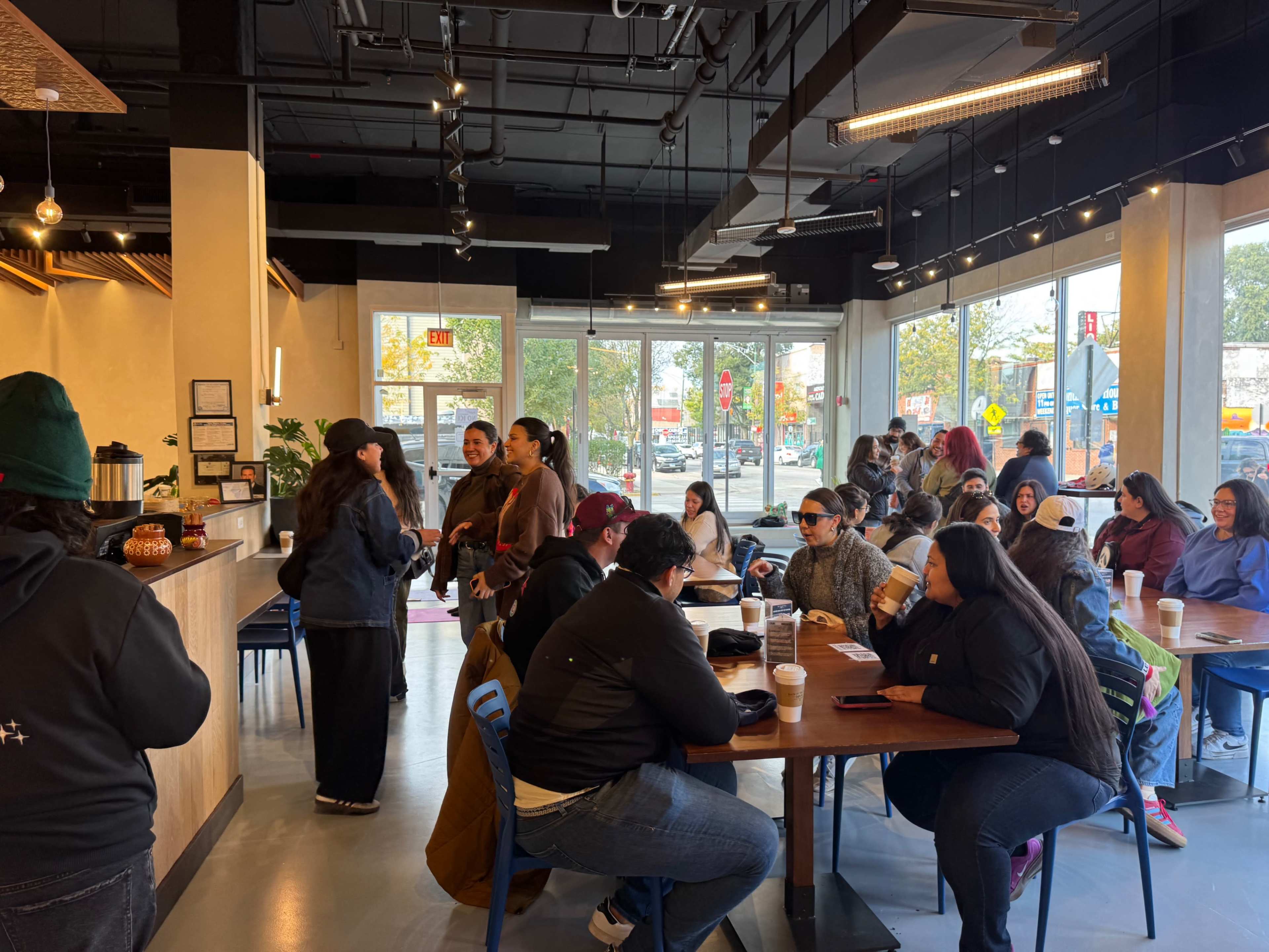 A crowded café features a mix of customers seated at tables and ordering at the counter, with large windows showing a bright, sunny day outside.