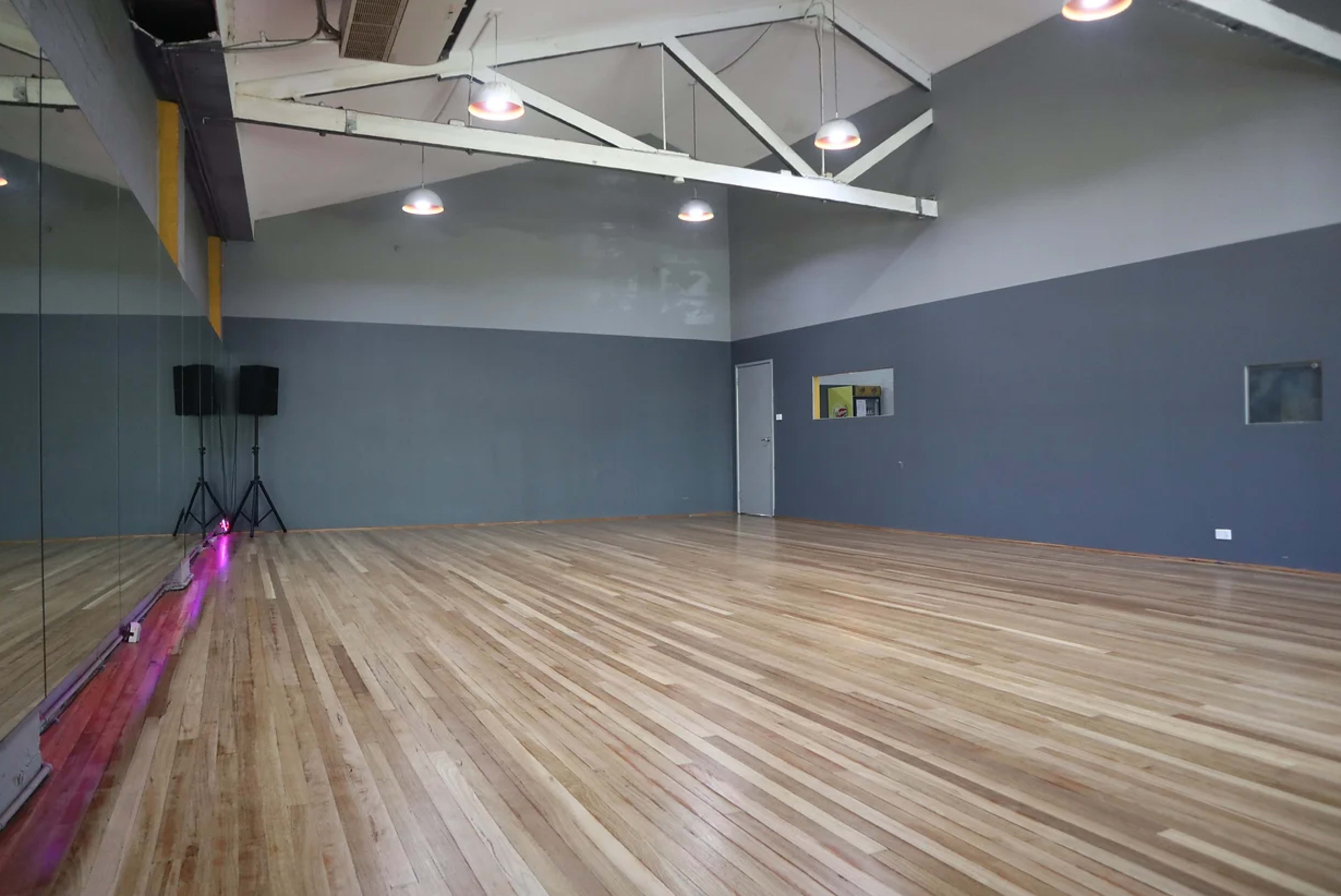 The image shows an empty dance studio with wooden flooring, gray walls, and large mirrors along one side.