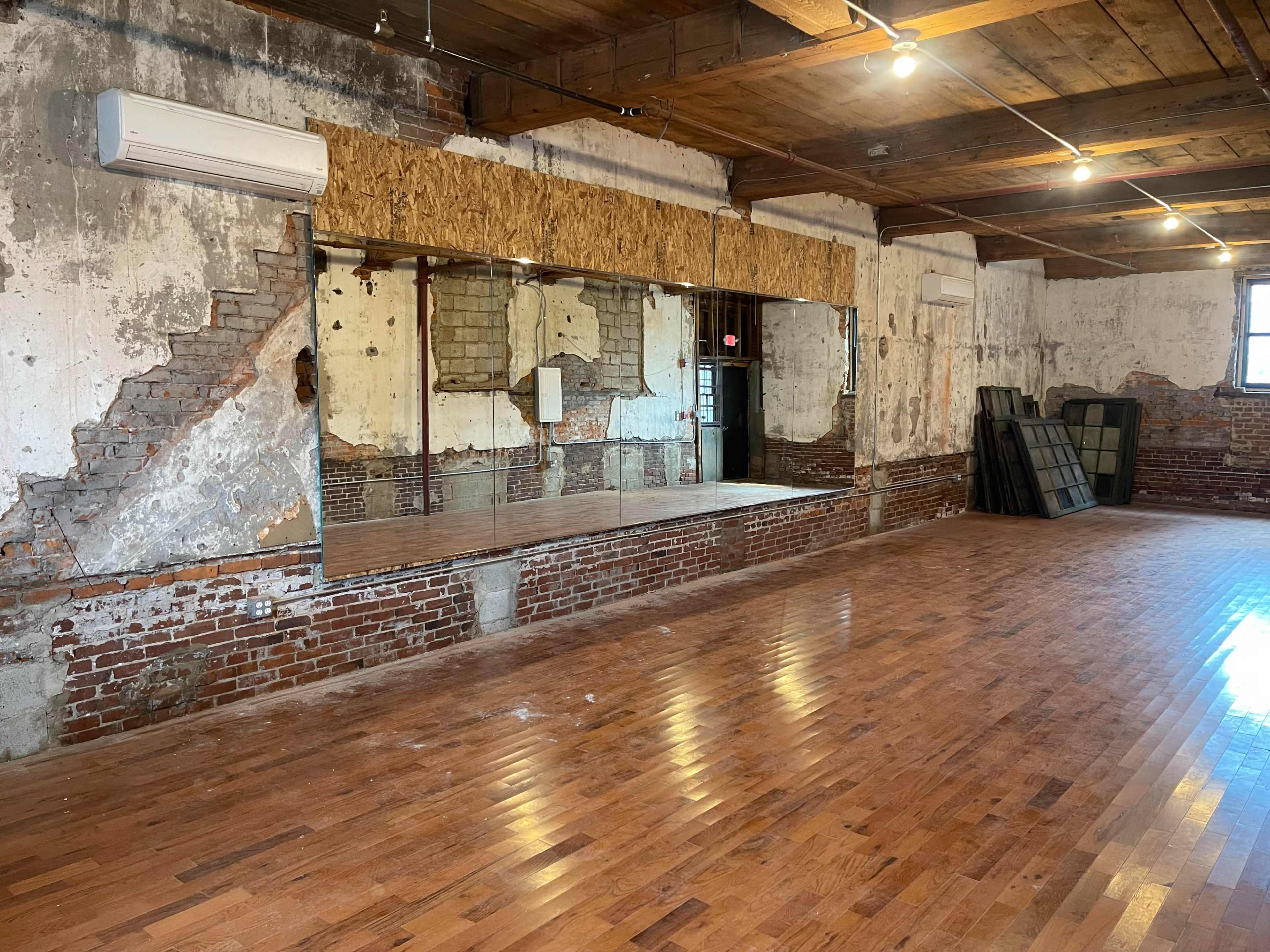 The image shows a spacious, wooden-floored room with exposed brick walls and a large mirror mounted on an unfinished wood panel.