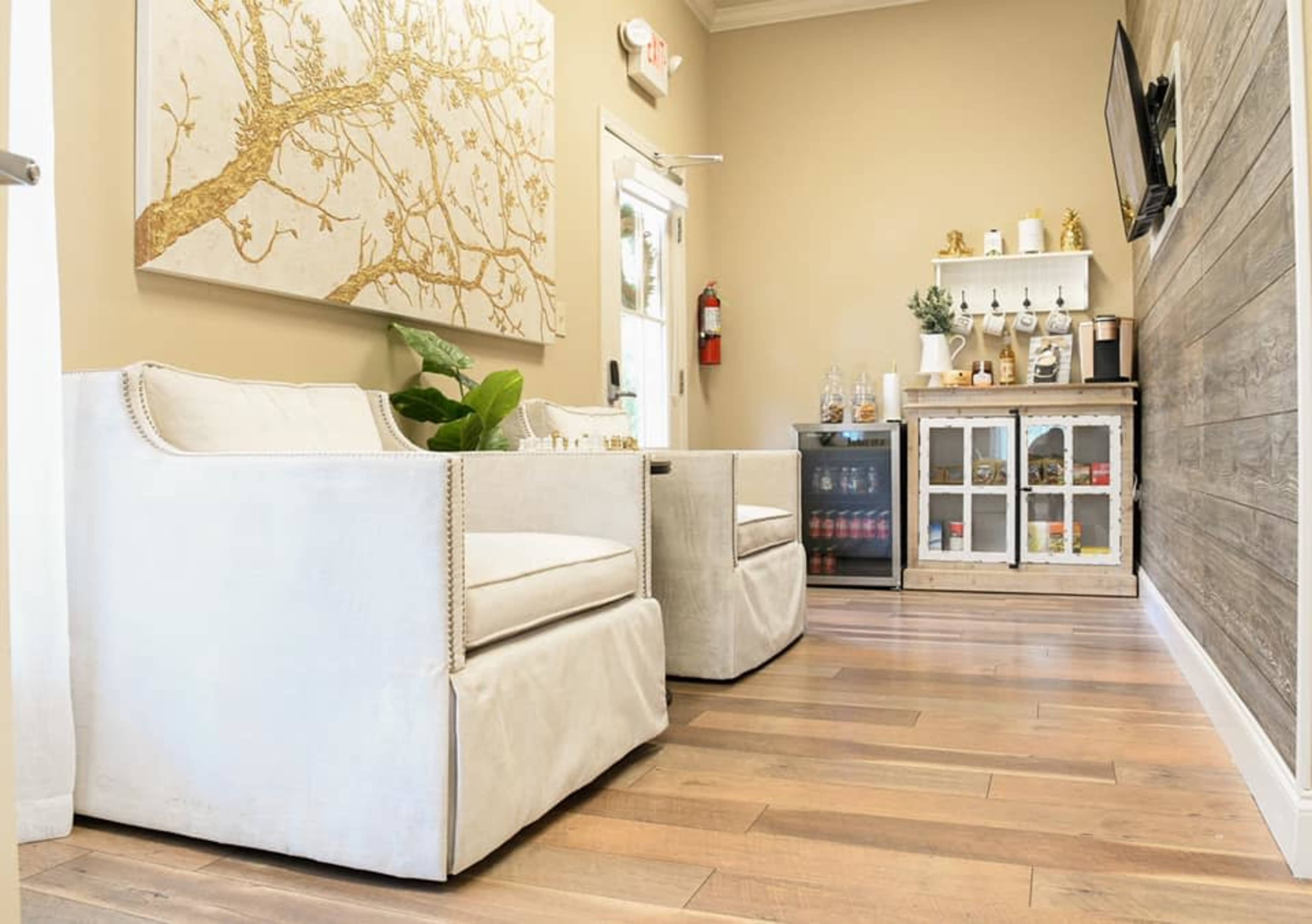 A well-lit interior space featuring two white armchairs, a small wooden cabinet with glass doors, and a decorative wall art piece, all set against wooden flooring.