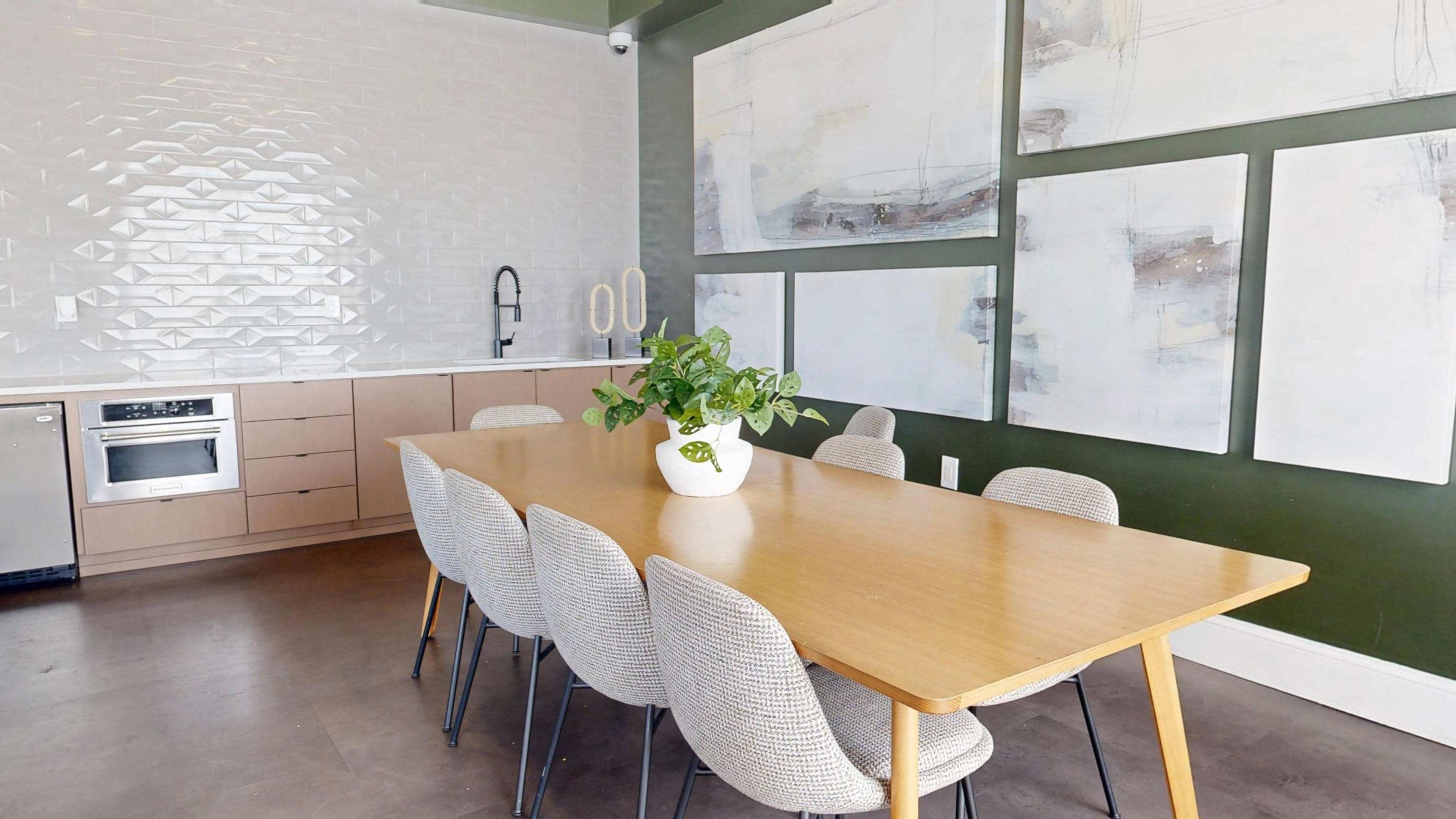 A dining area features a long wooden table surrounded by upholstered chairs, with modern artwork and a textured backsplash on the walls.