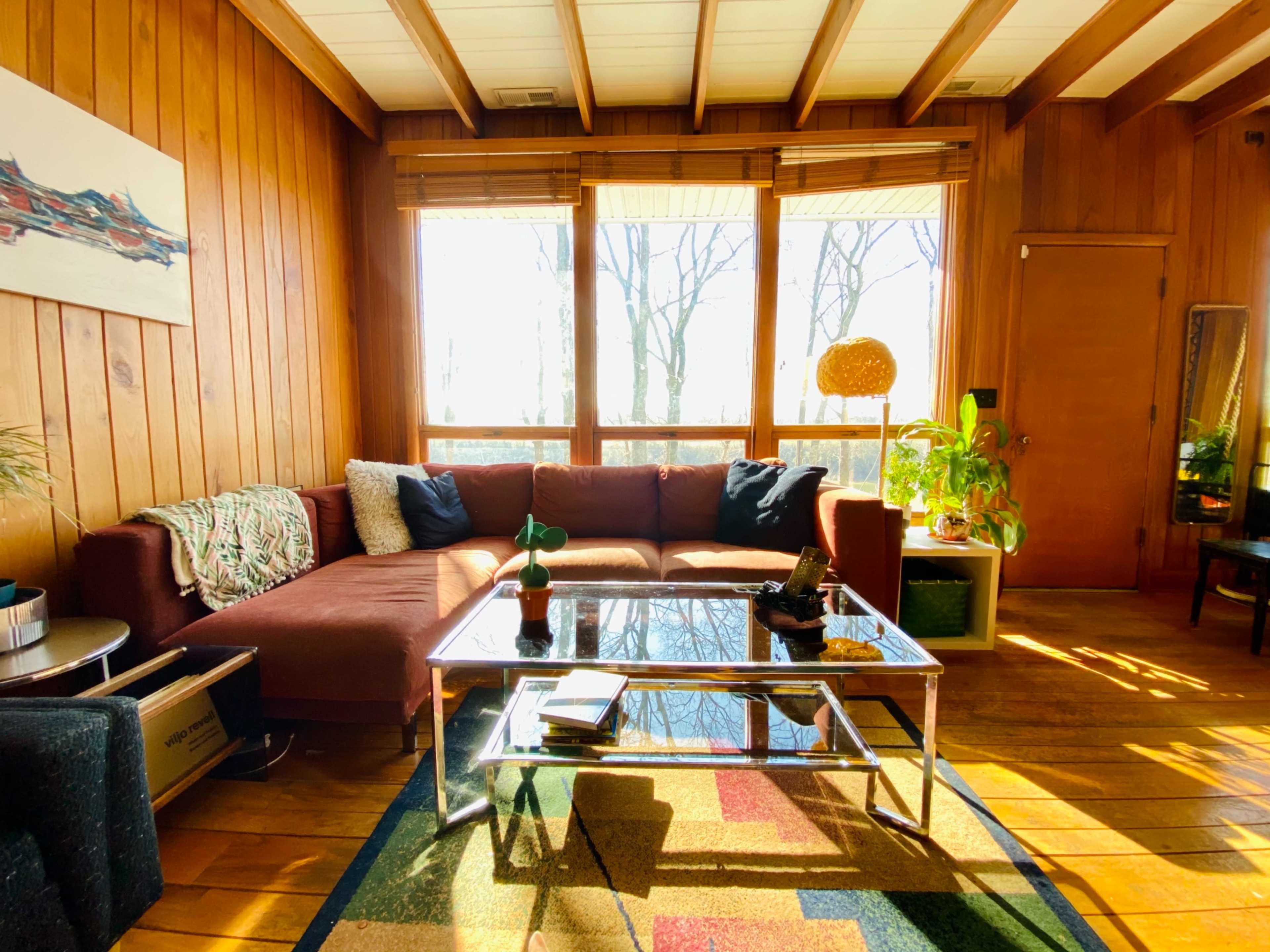 A cozy living room features a large brown couch, a glass coffee table, and sunlight streaming through windows with wooden walls and a colorful rug.