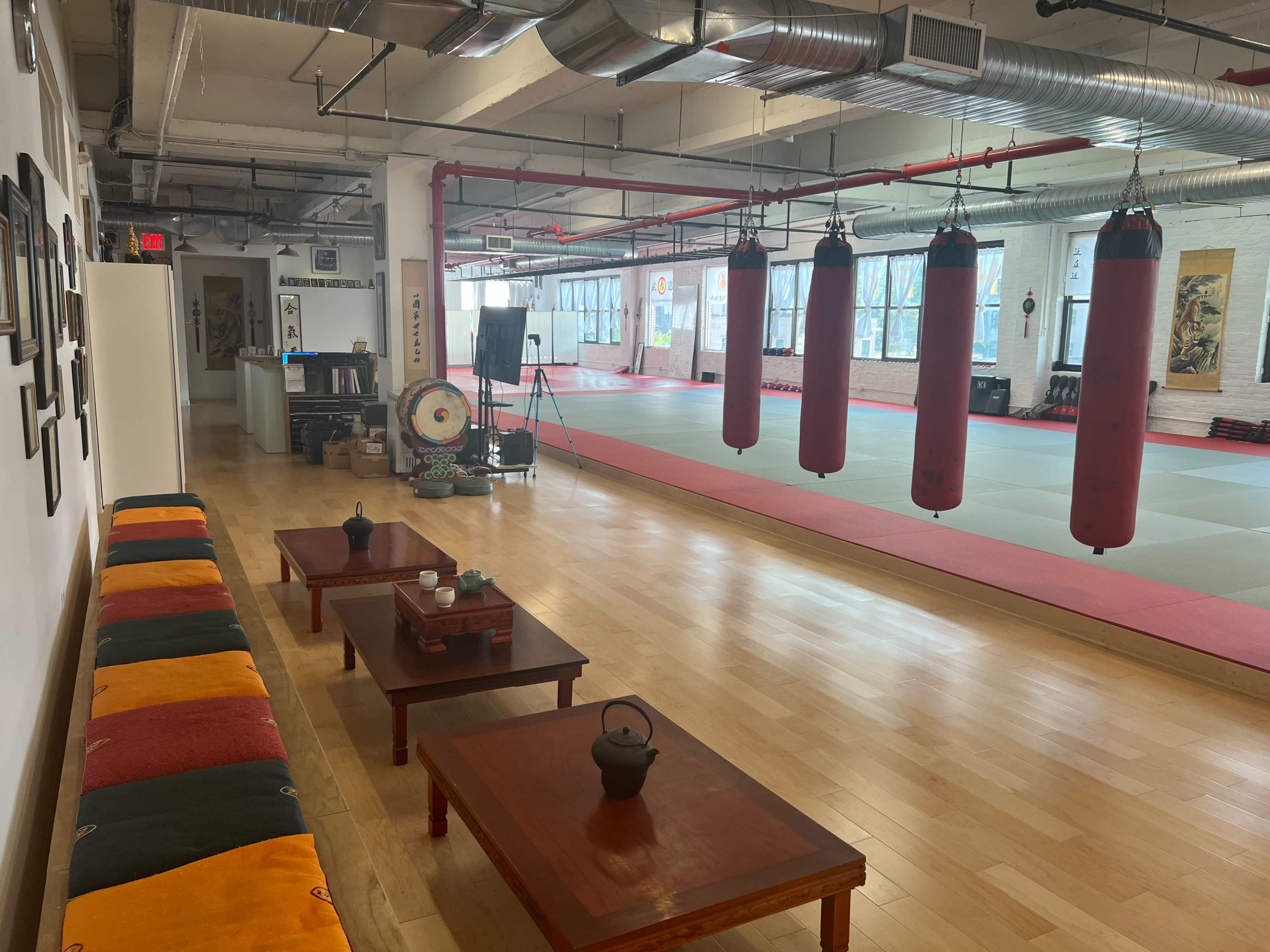 A spacious martial arts studio with hanging punching bags, wooden tables, and a seating area along the wall.