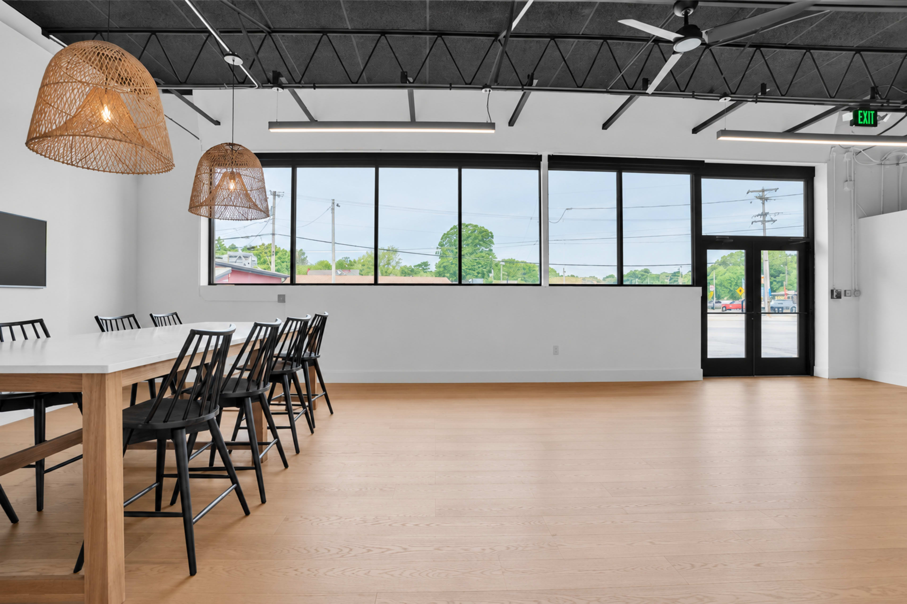The image shows a spacious, modern room with a long table and black chairs, featuring large windows that allow natural light to fill the space.