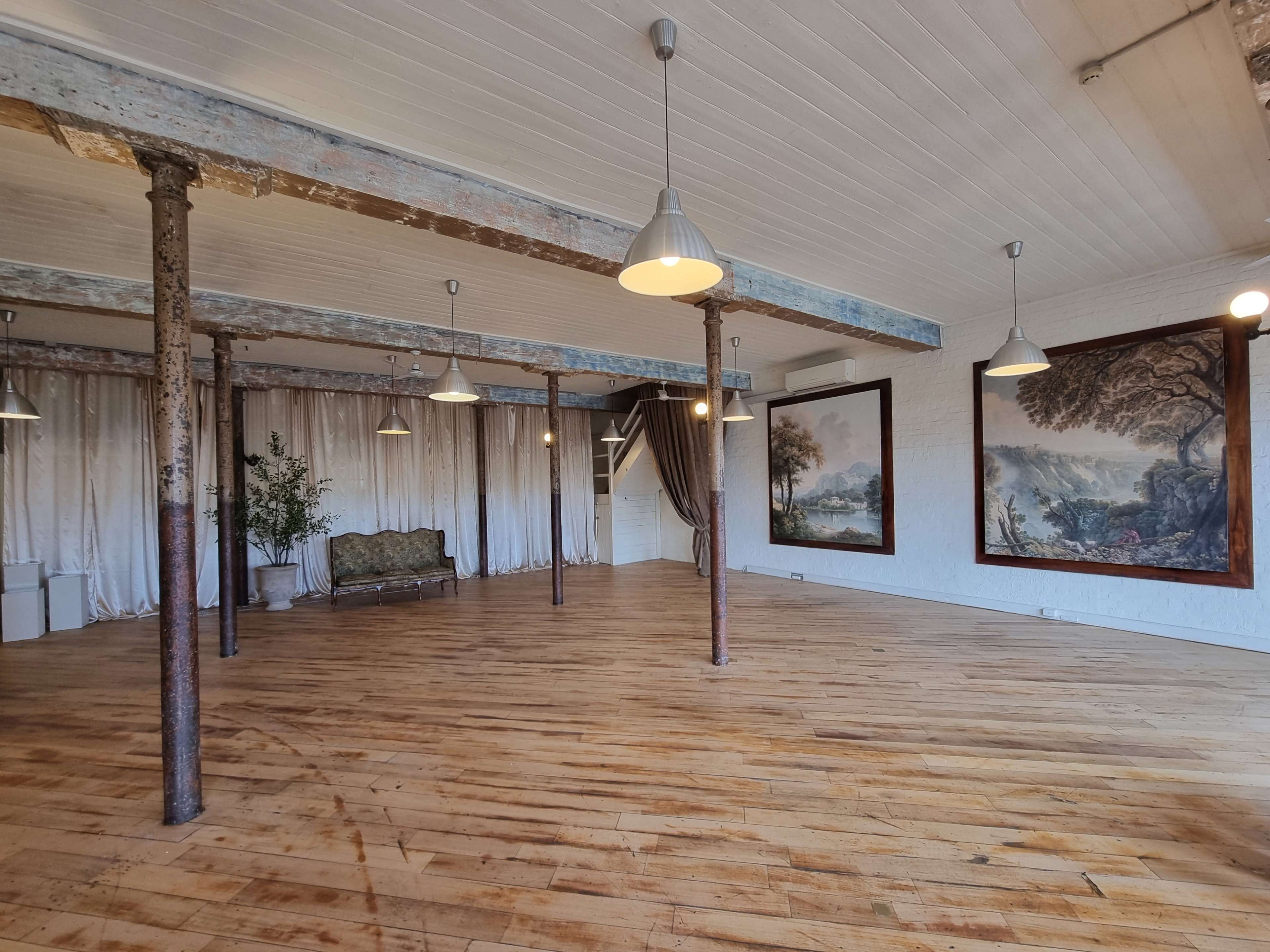 The image features a spacious room with wooden flooring, exposed metal columns, and large framed paintings on the white walls, illuminated by hanging lights.