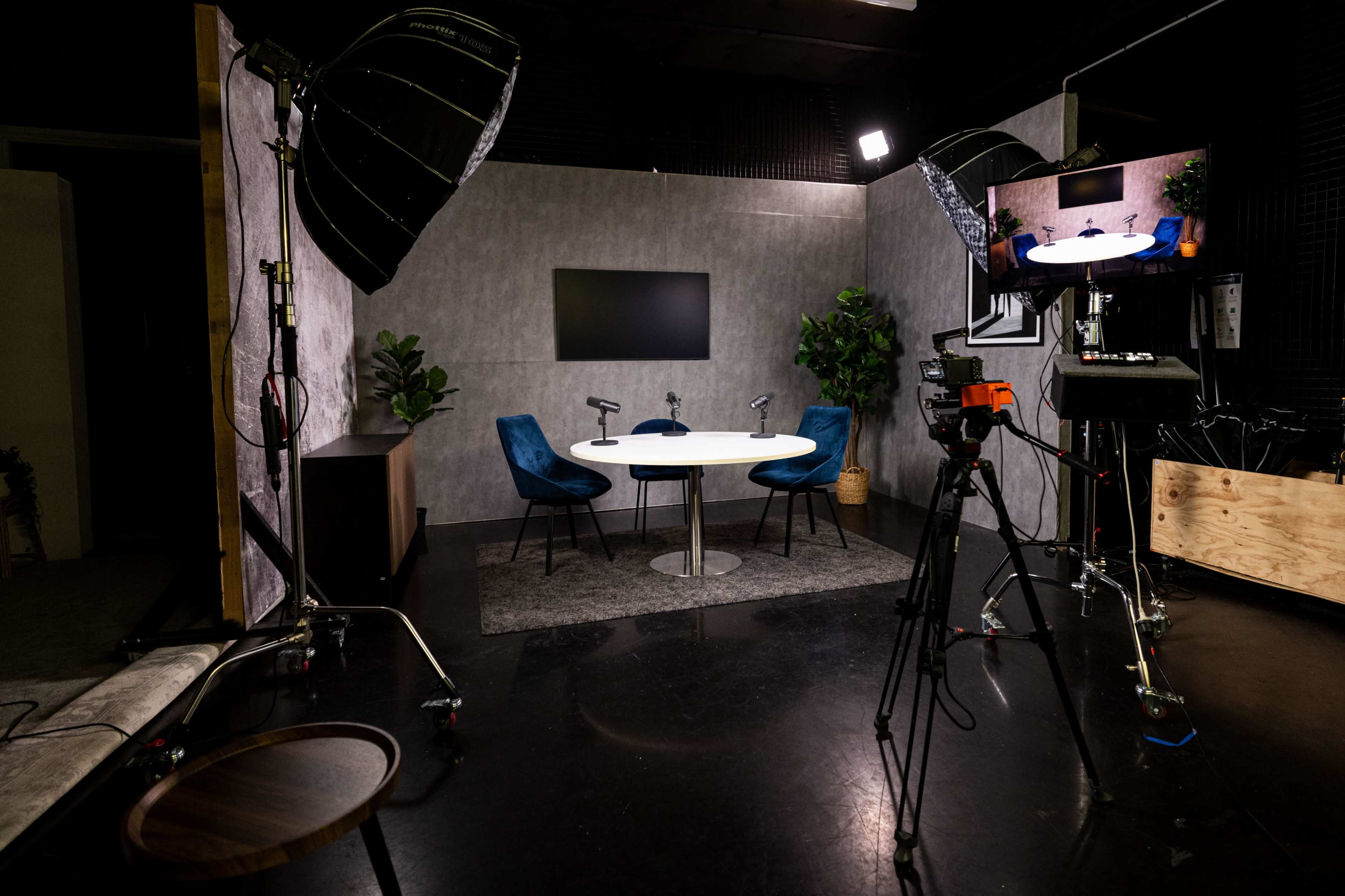 The image shows a modern studio setup with a round table, two blue chairs, a television screen on the wall, and camera equipment ready for recording.