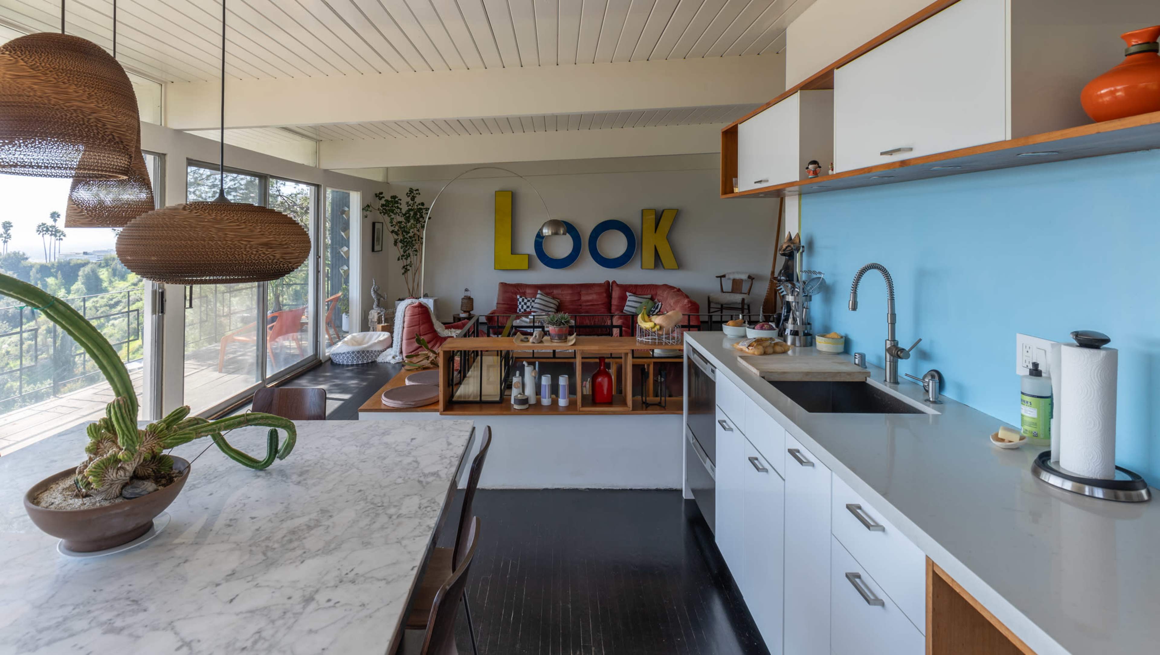 A modern kitchen features a marble countertop, sleek cabinets, and an open view into a living area with colorful decor and a red sofa.