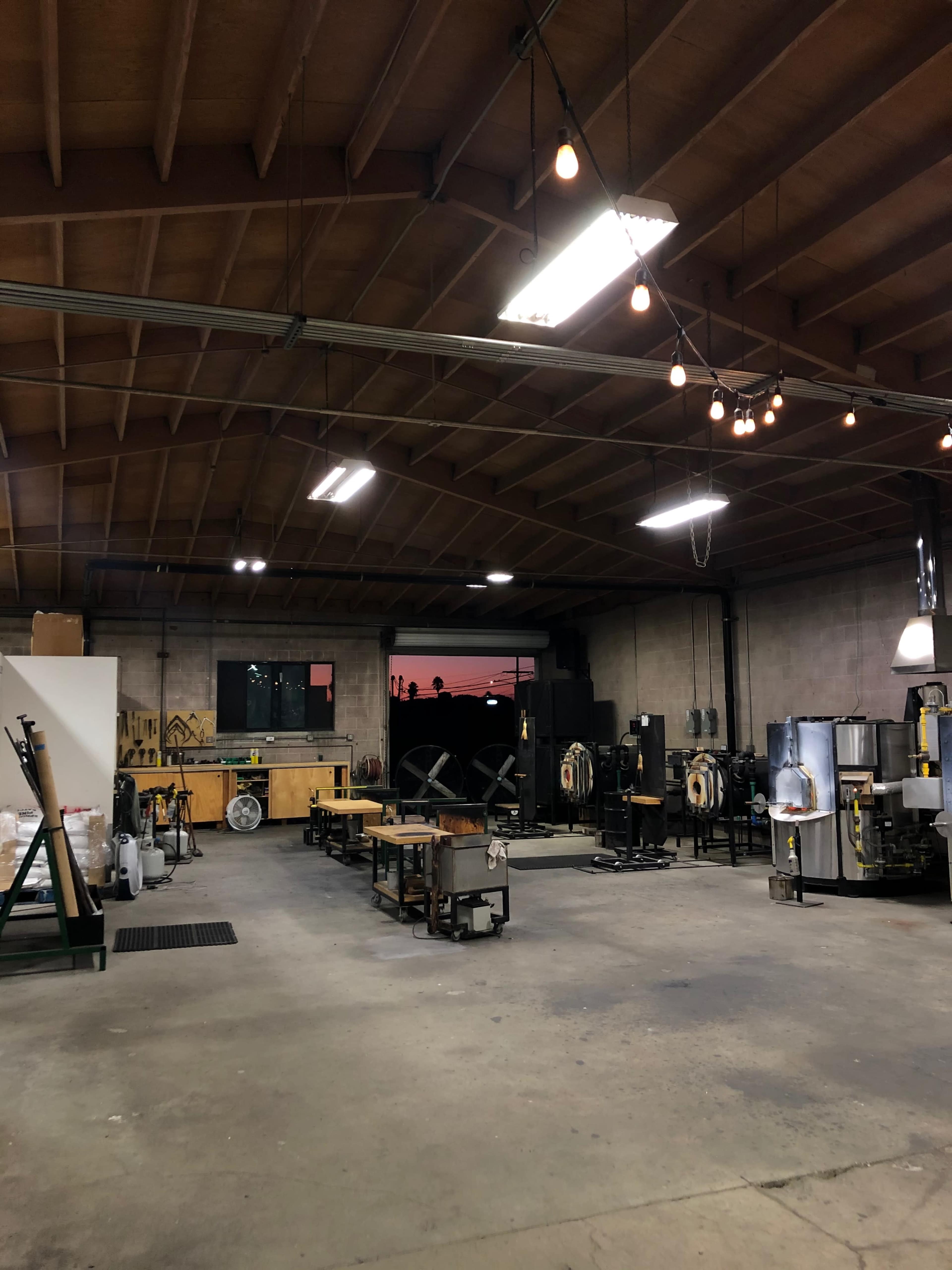 The image shows an industrial workshop with various equipment, tables, and lighting fixtures, featuring a view of a colorful sunset through a large window.