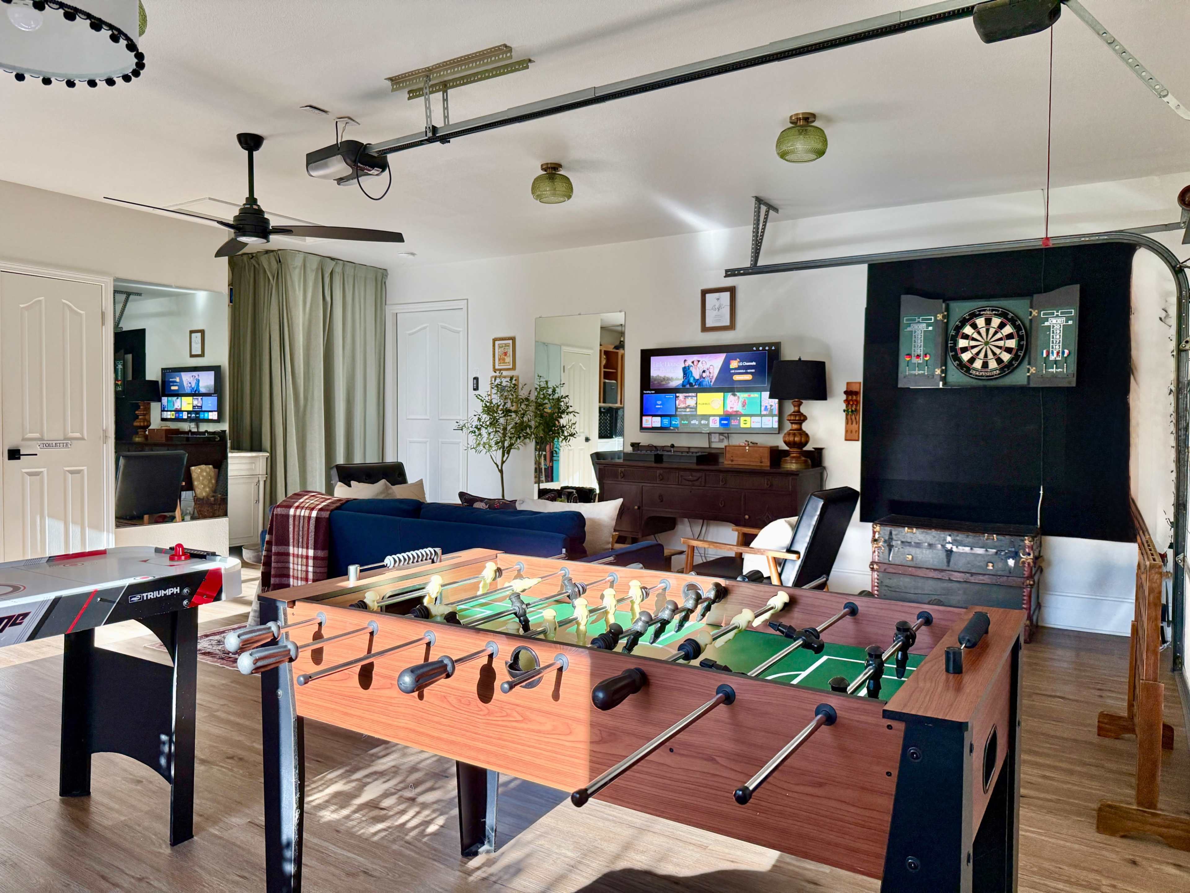 The image shows a recreational room featuring a foosball table, a dartboard, two televisions, and a cozy sitting area with a couch and chairs.