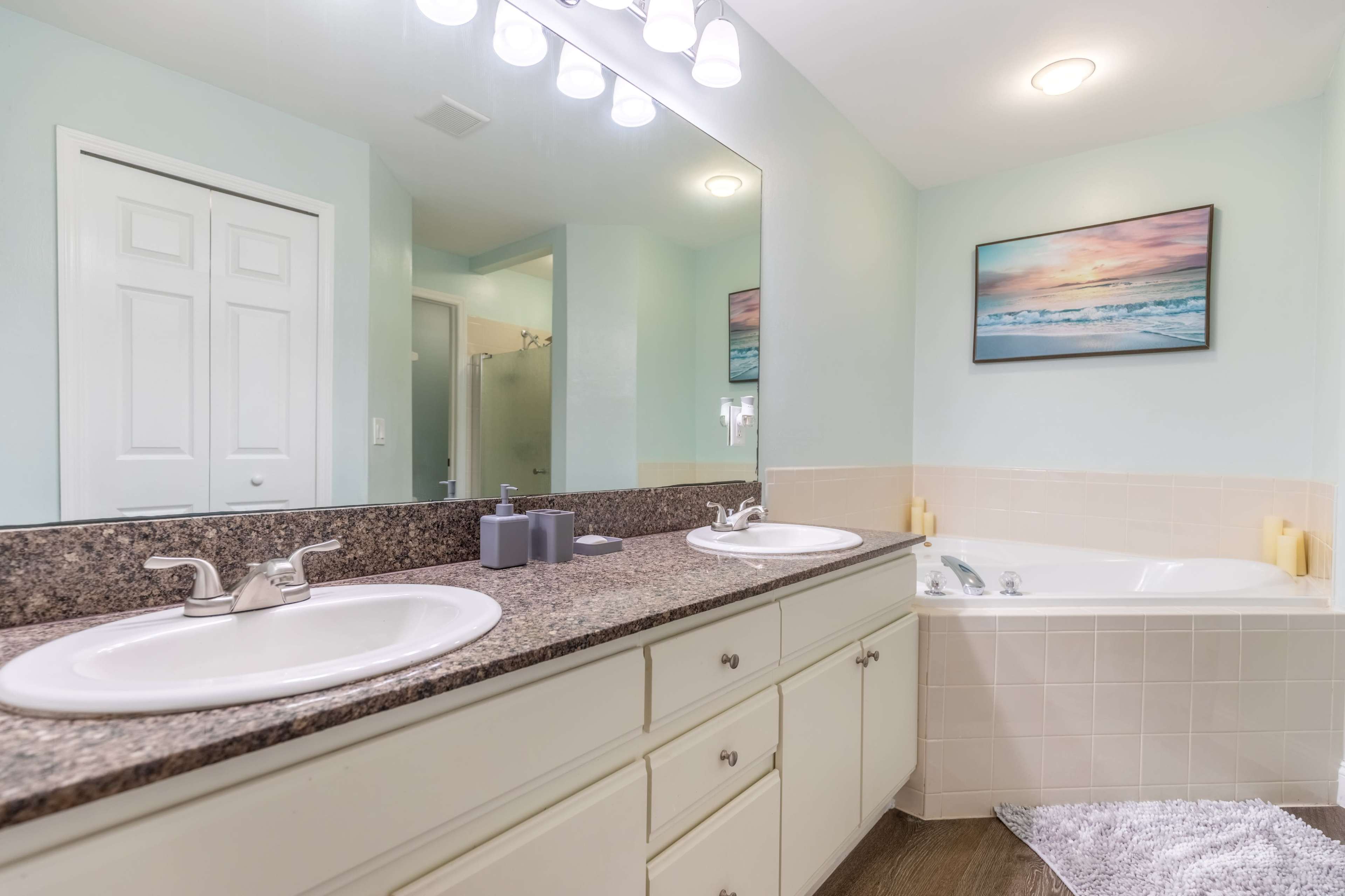 A bathroom with double sinks, a bathtub, and a mirrored vanity, set against light blue walls and a beach-themed painting.