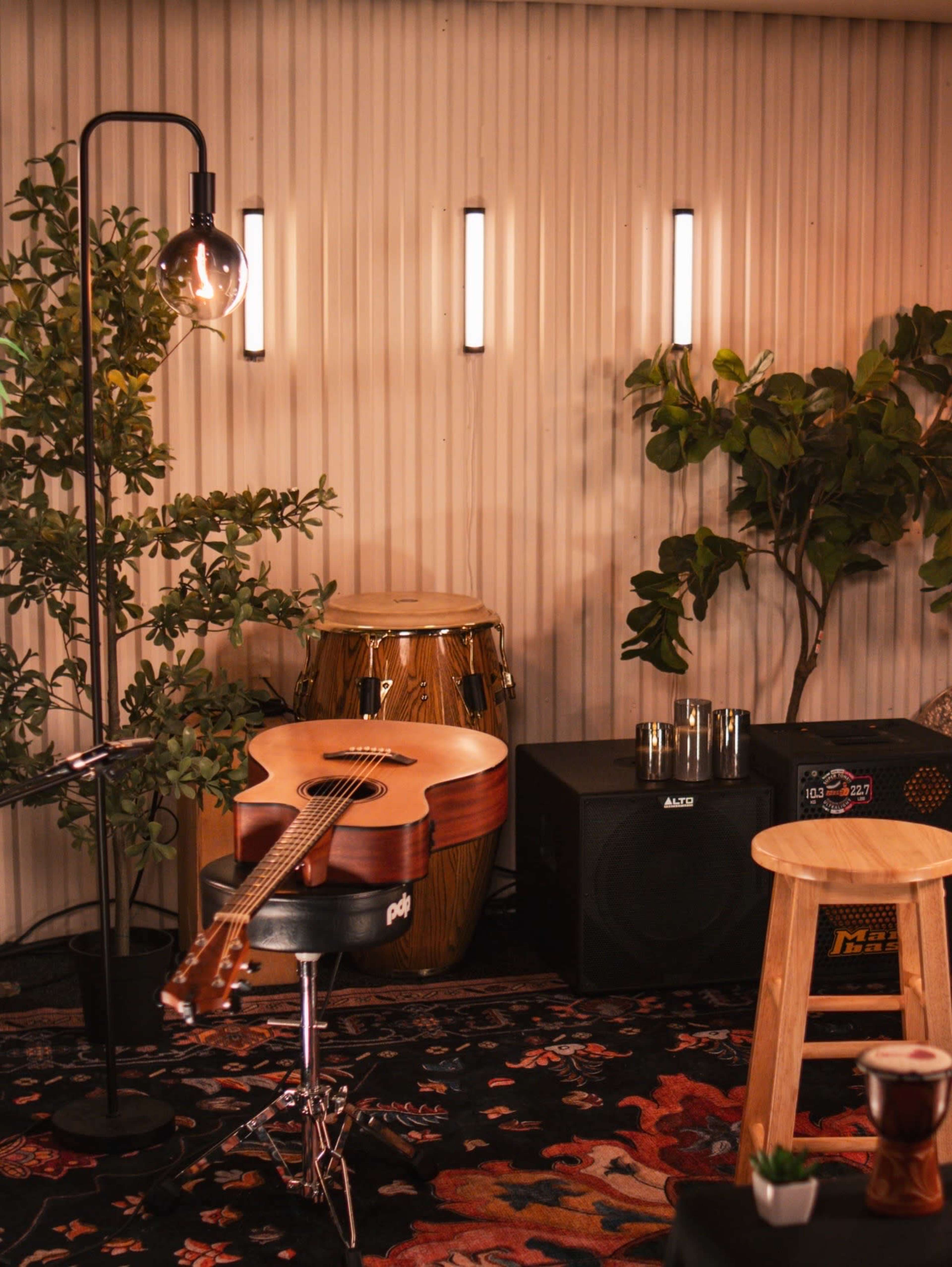 The image shows a cozy music room with an acoustic guitar resting on a stool, conga drums in the background, and a speaker beside a tall plant and a floor lamp.