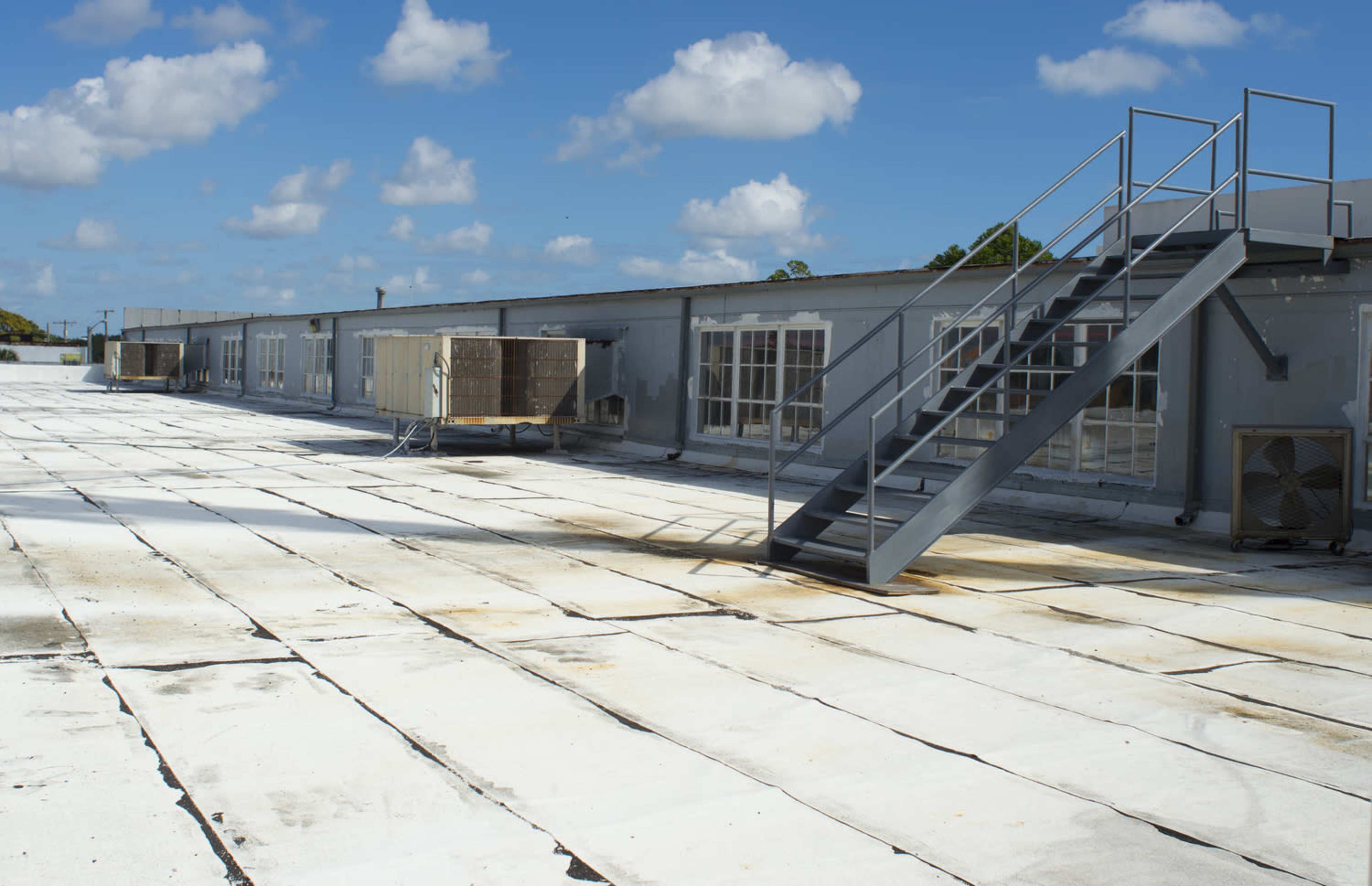A flat rooftop features a metal staircase, air conditioning units, and scattered clouds in the blue sky above.