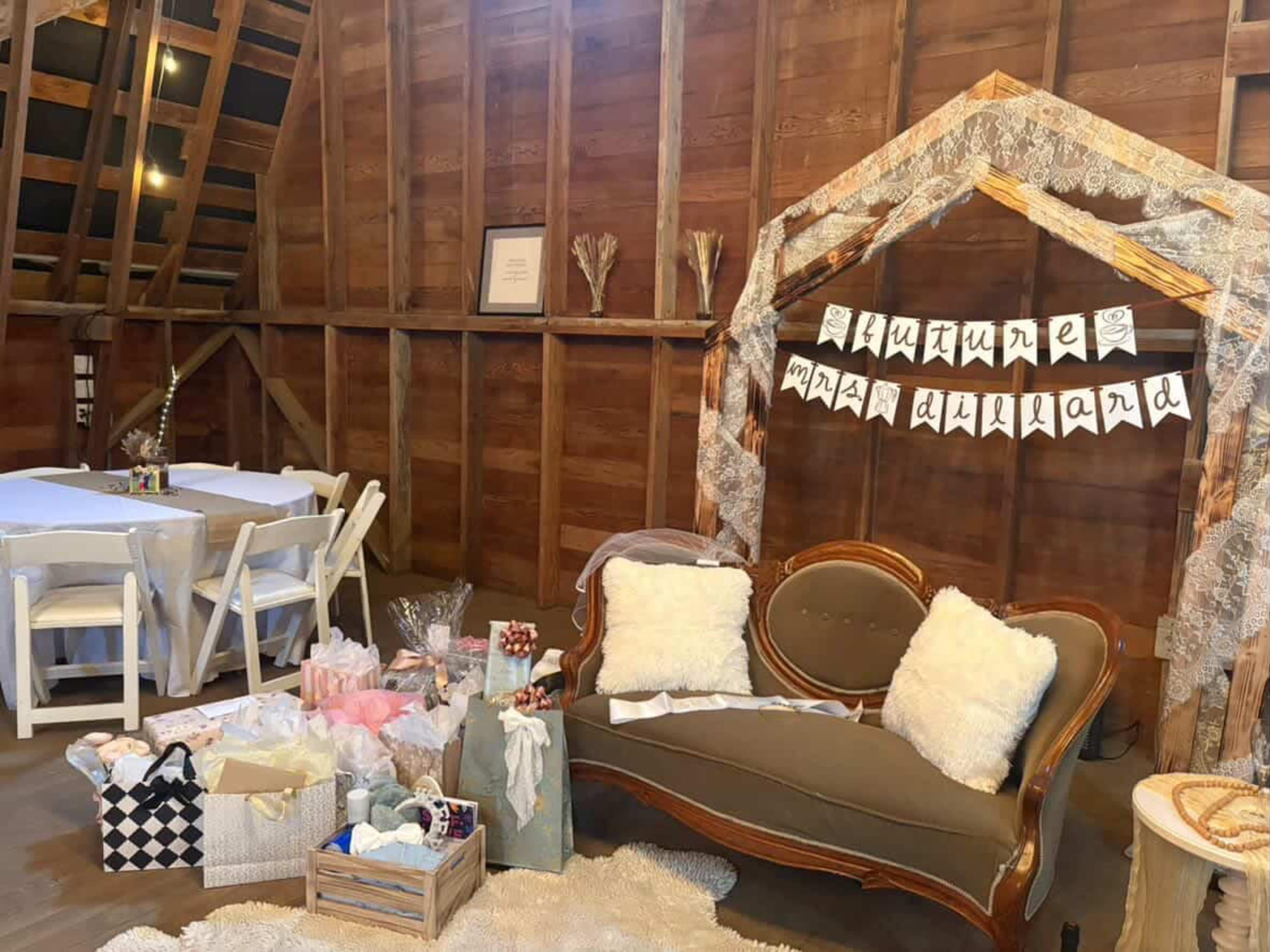 The image shows a rustic interior setting featuring a vintage sofa, a decorated archway with a banner, and neatly arranged tables, all surrounded by wooden walls.