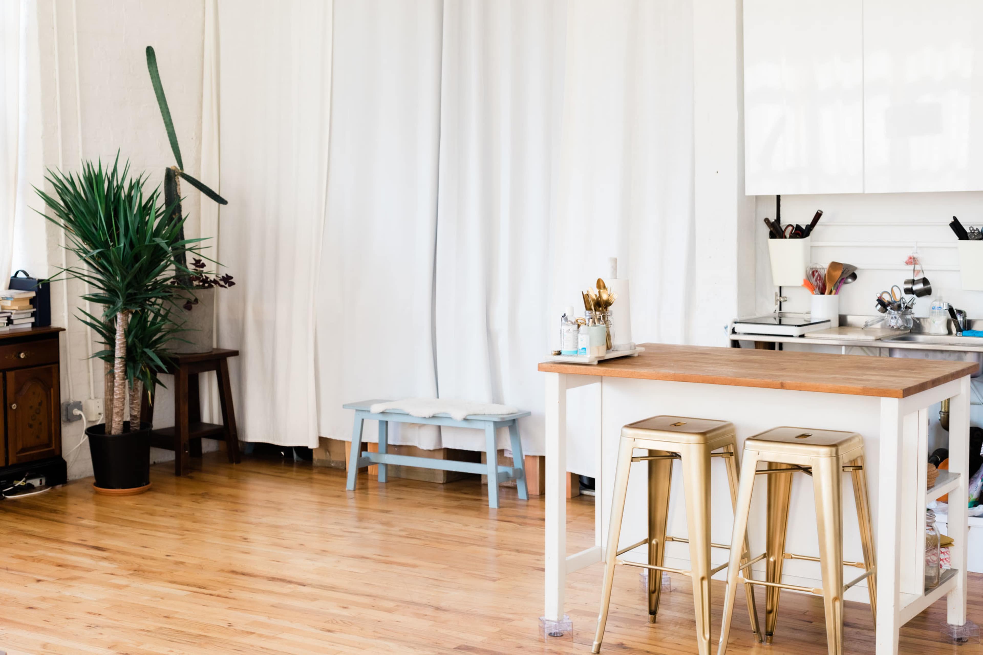 A kitchen space features a wooden island with two metal stools, plants, and a curtain separating the area from the rest of the room.
