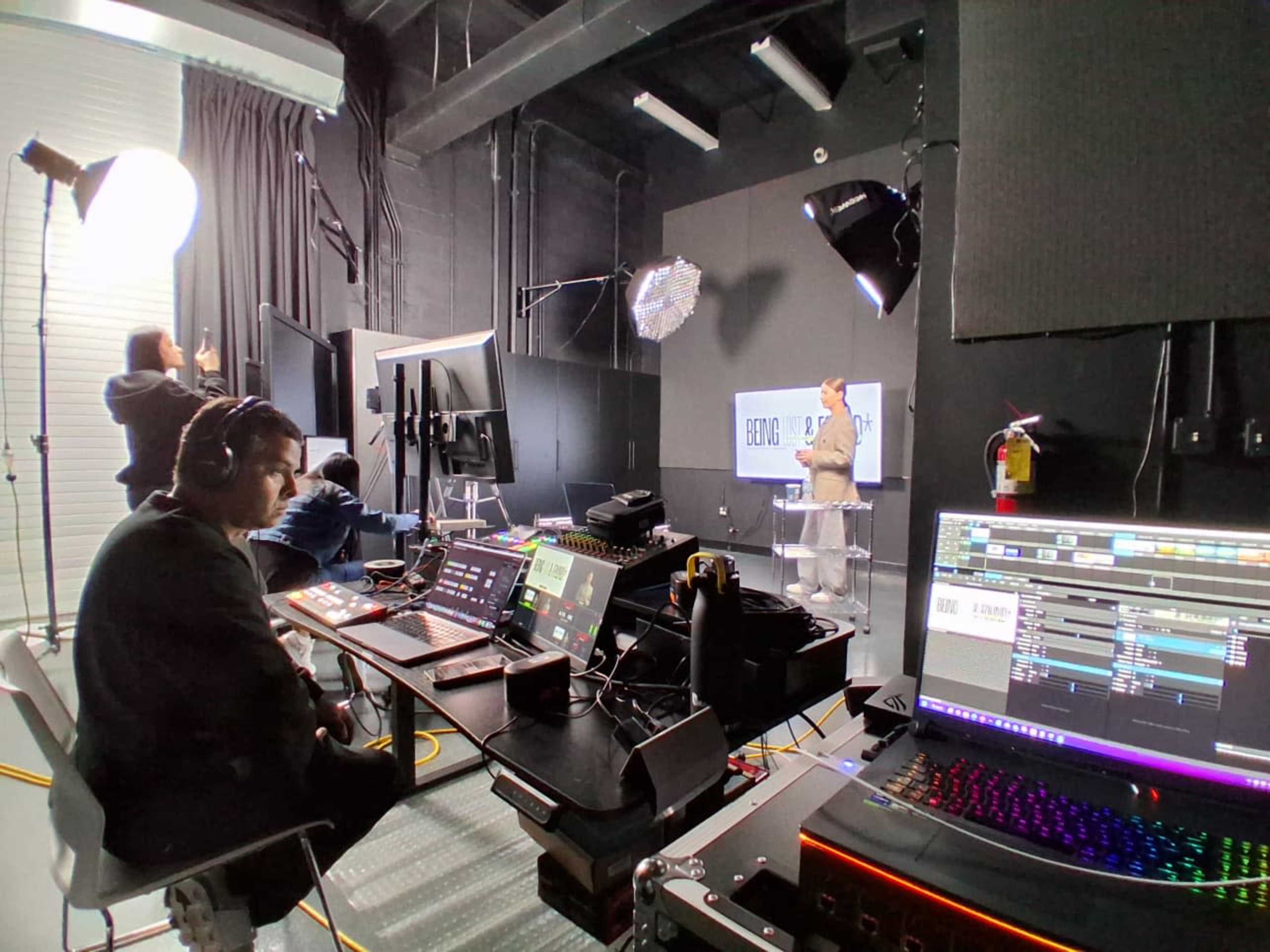 A group of people are working in a studio setting, with one person presenting at a podium while others operate technical equipment at a control station.