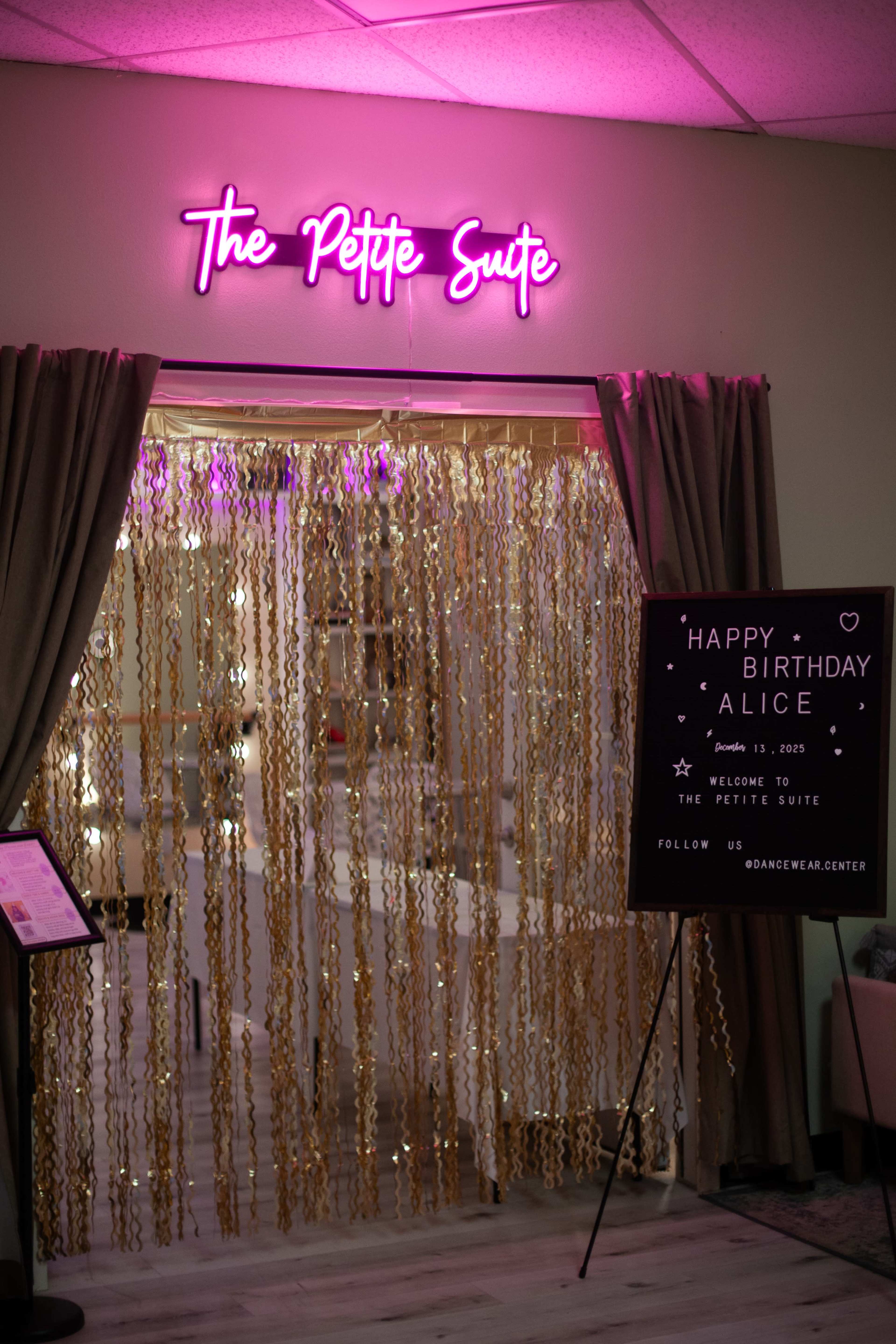 The image shows a decorated entrance to a space called "The Petite Suite," featuring a neon sign, a curtain made of golden strands, and a welcome sign for a birthday celebration.