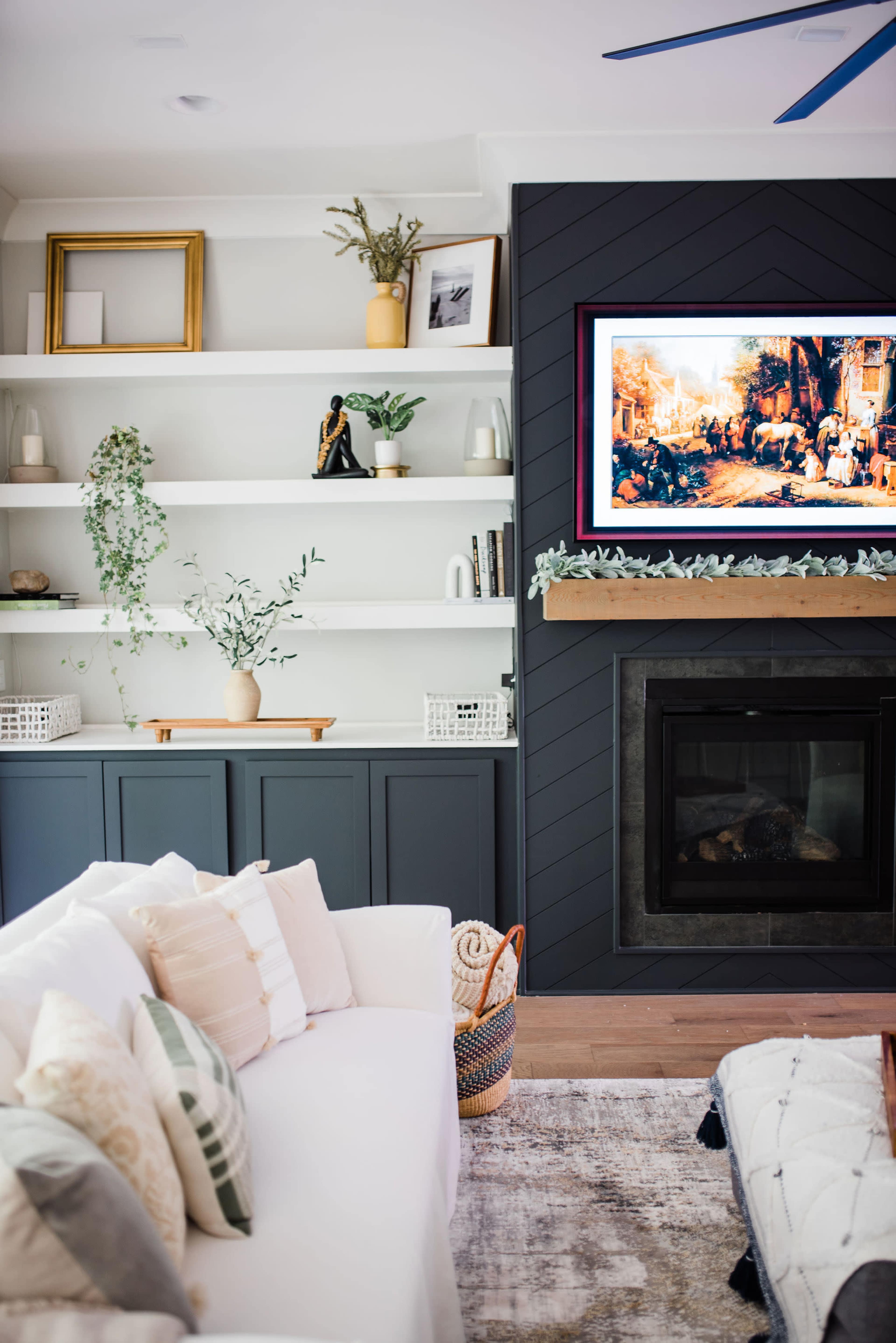 A modern living room features a white sofa, a dark accent wall with a television above a fireplace, and built-in shelves displaying decorative items and plants.