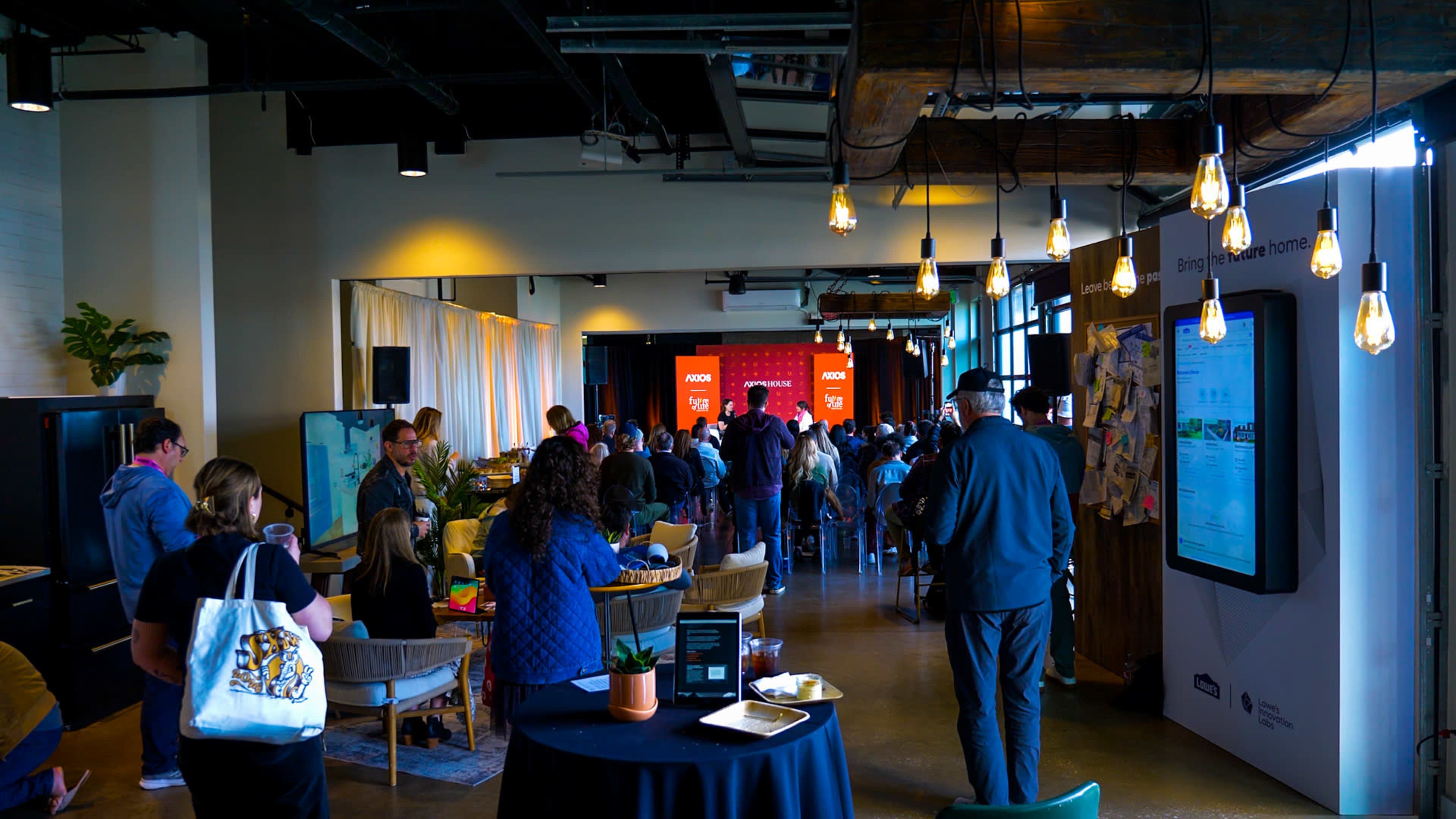 A crowd gathers in a large, well-lit room for an event featuring a presentation backdrop.