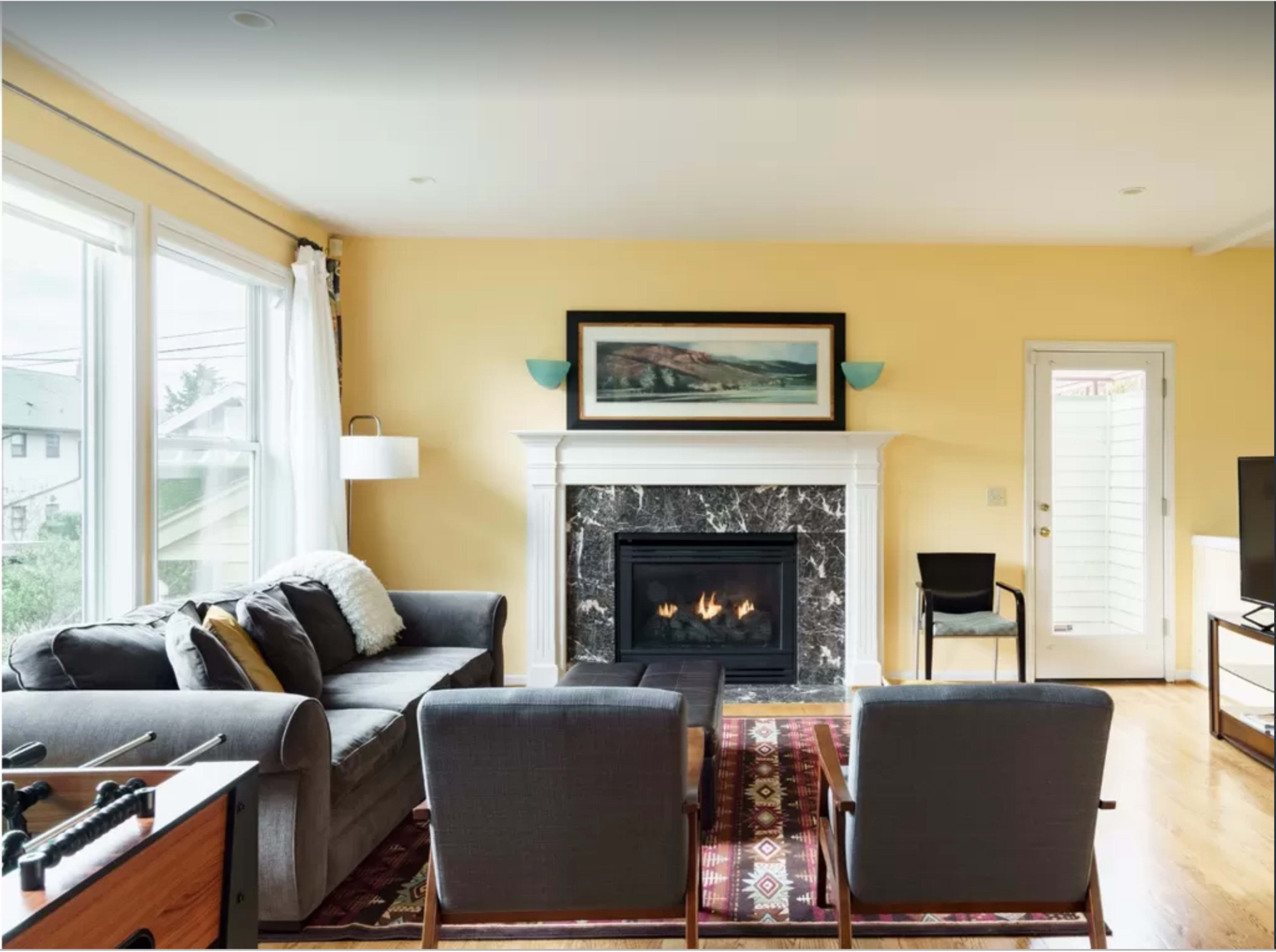 The room features a gray sofa, two armchairs, a fireplace with a marble surround, and a door leading outside, with natural light coming through large windows.