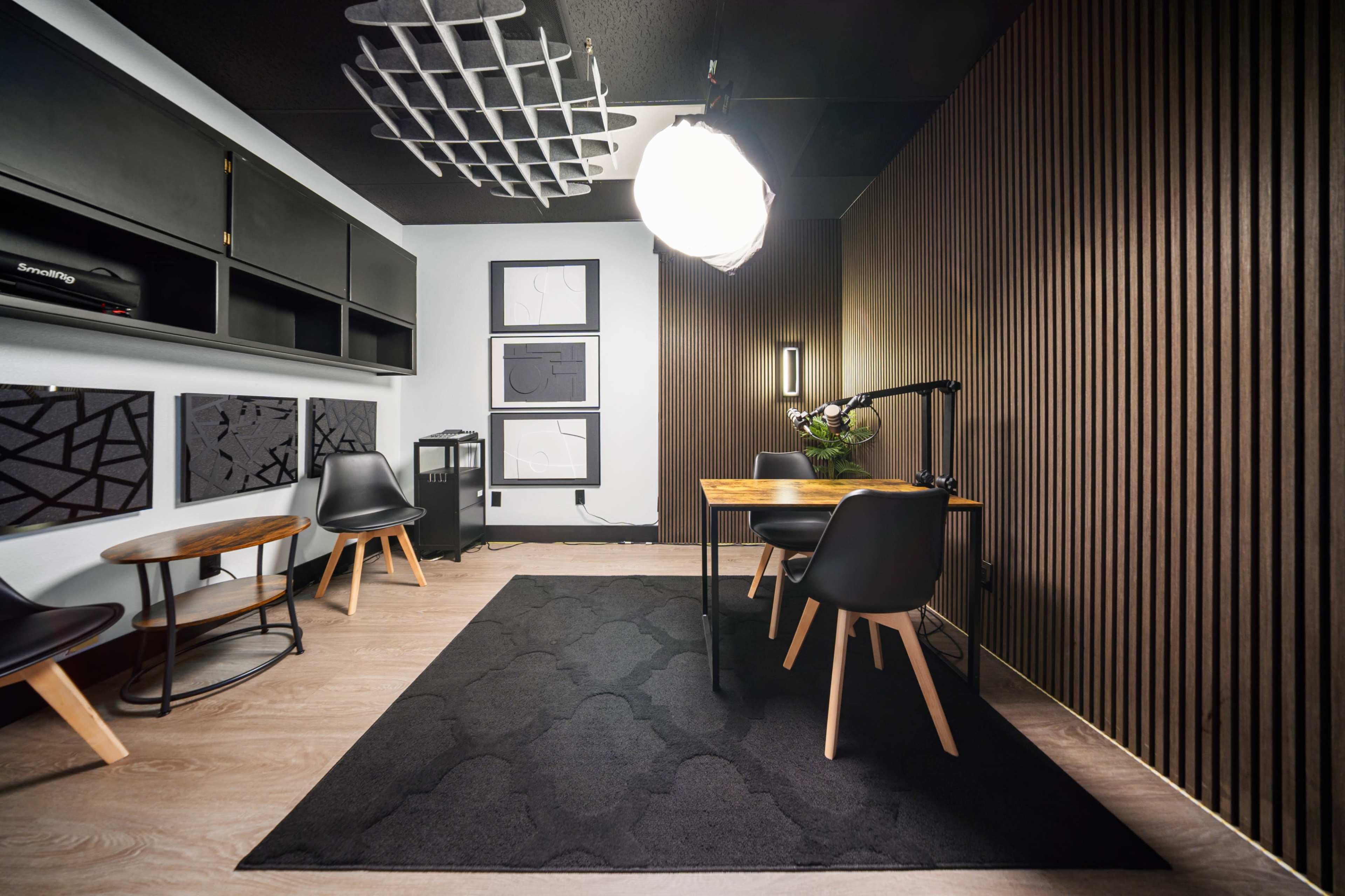 The image shows a modern studio space with a wooden table and black chairs, accented by a textured wall and soft lighting.