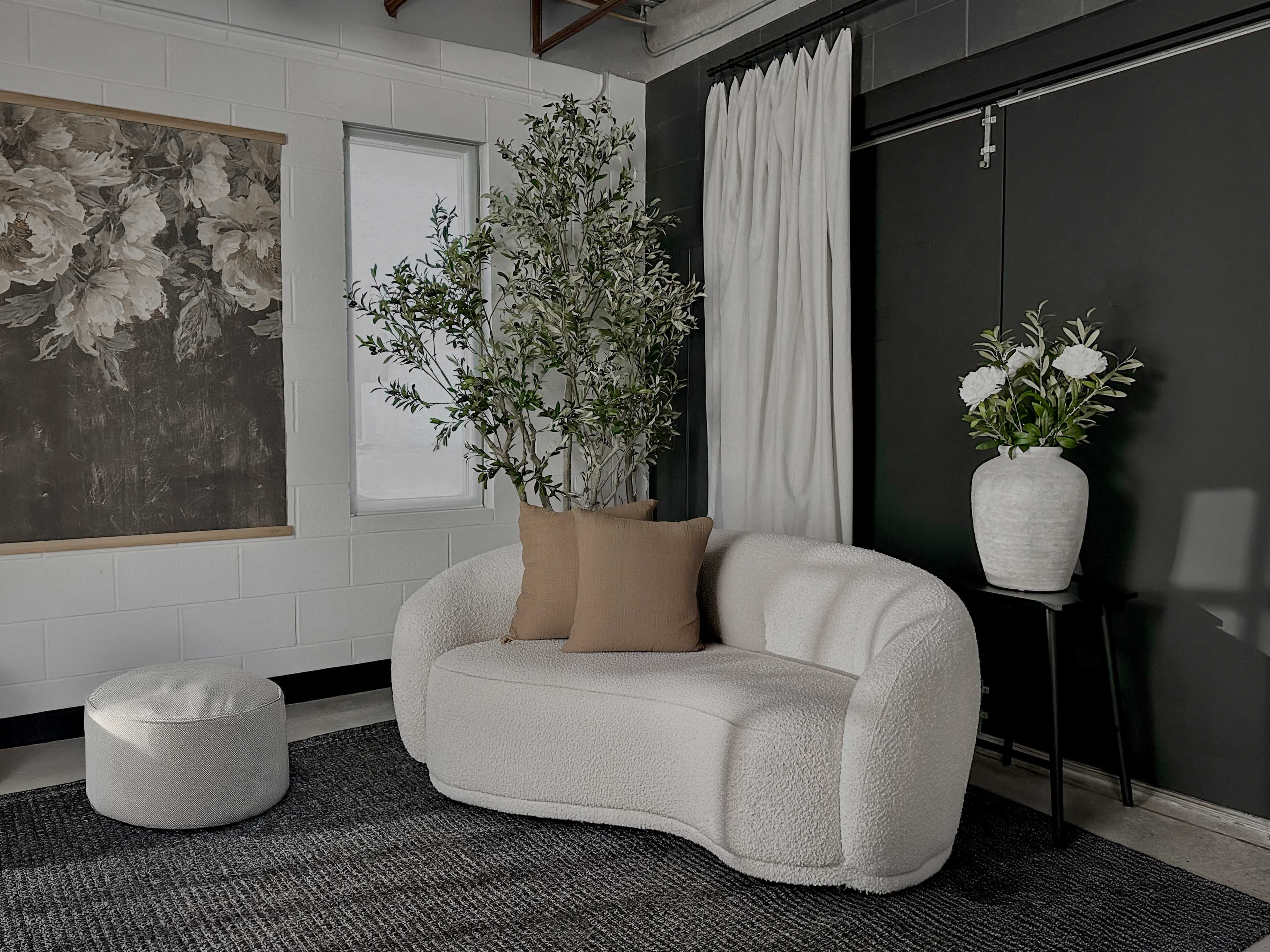 The image shows a cozy seating area featuring a curved white sofa with a beige pillow, a small round ottoman, and a black wall with a floral artwork and potted plants.