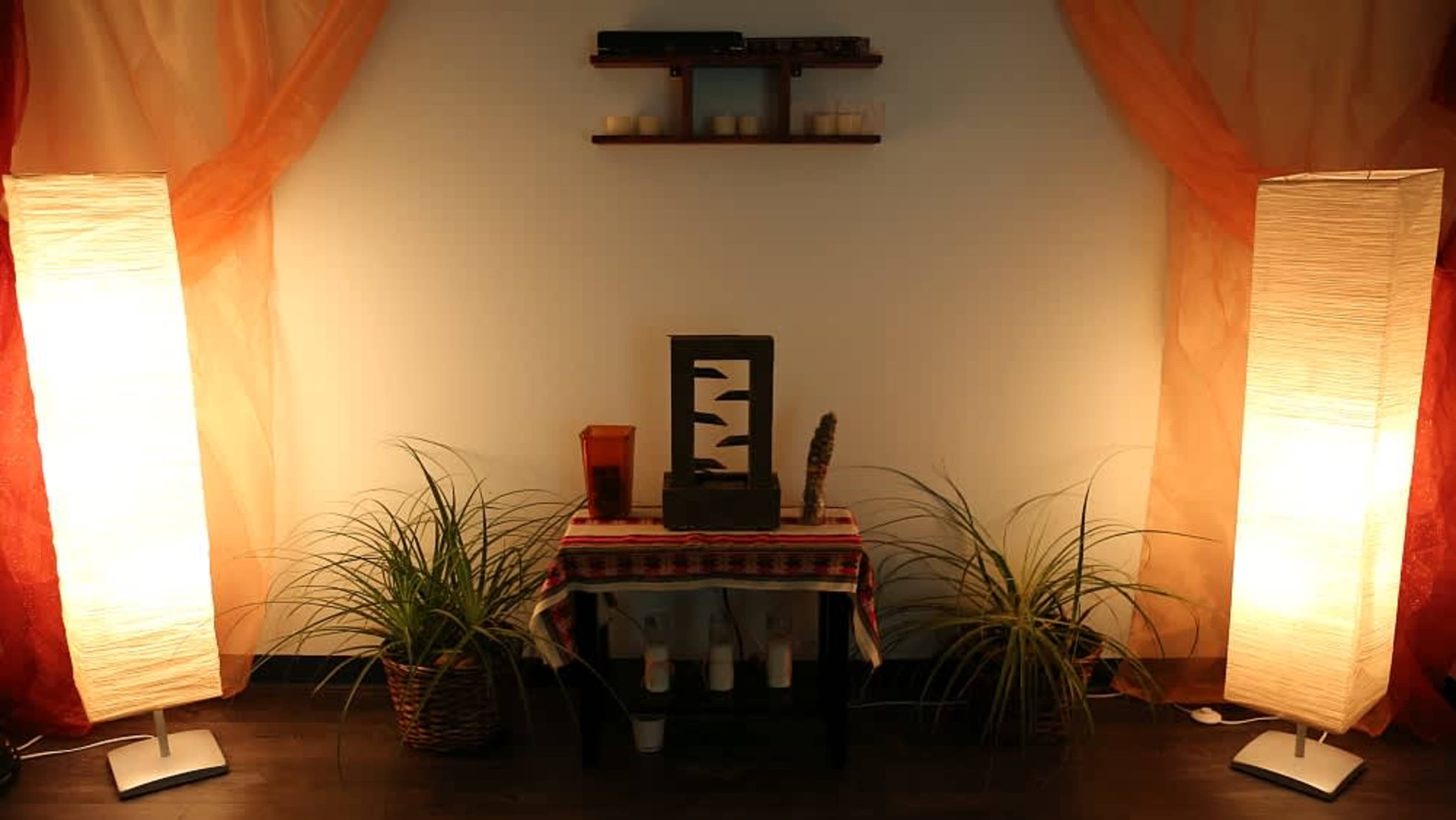 The image features a tranquil indoor setting with two illuminated floor lamps, a decorative table with a small fountain, and potted plants, all framed by sheer drapes.