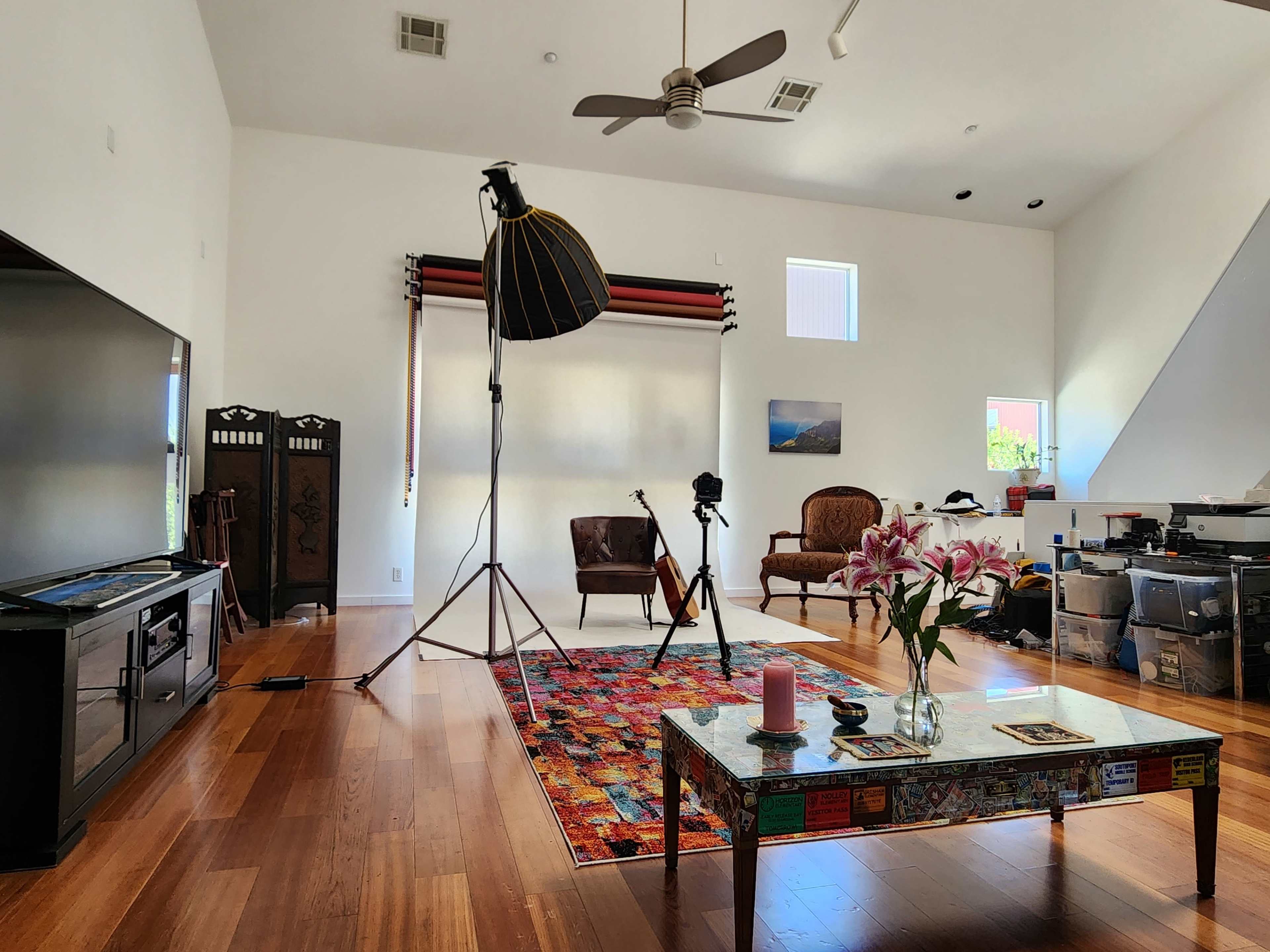 A spacious, well-lit living room with a colorful rug, a large television, a vintage-style armchair, a photography setup, and a coffee table adorned with flowers and decorative items.