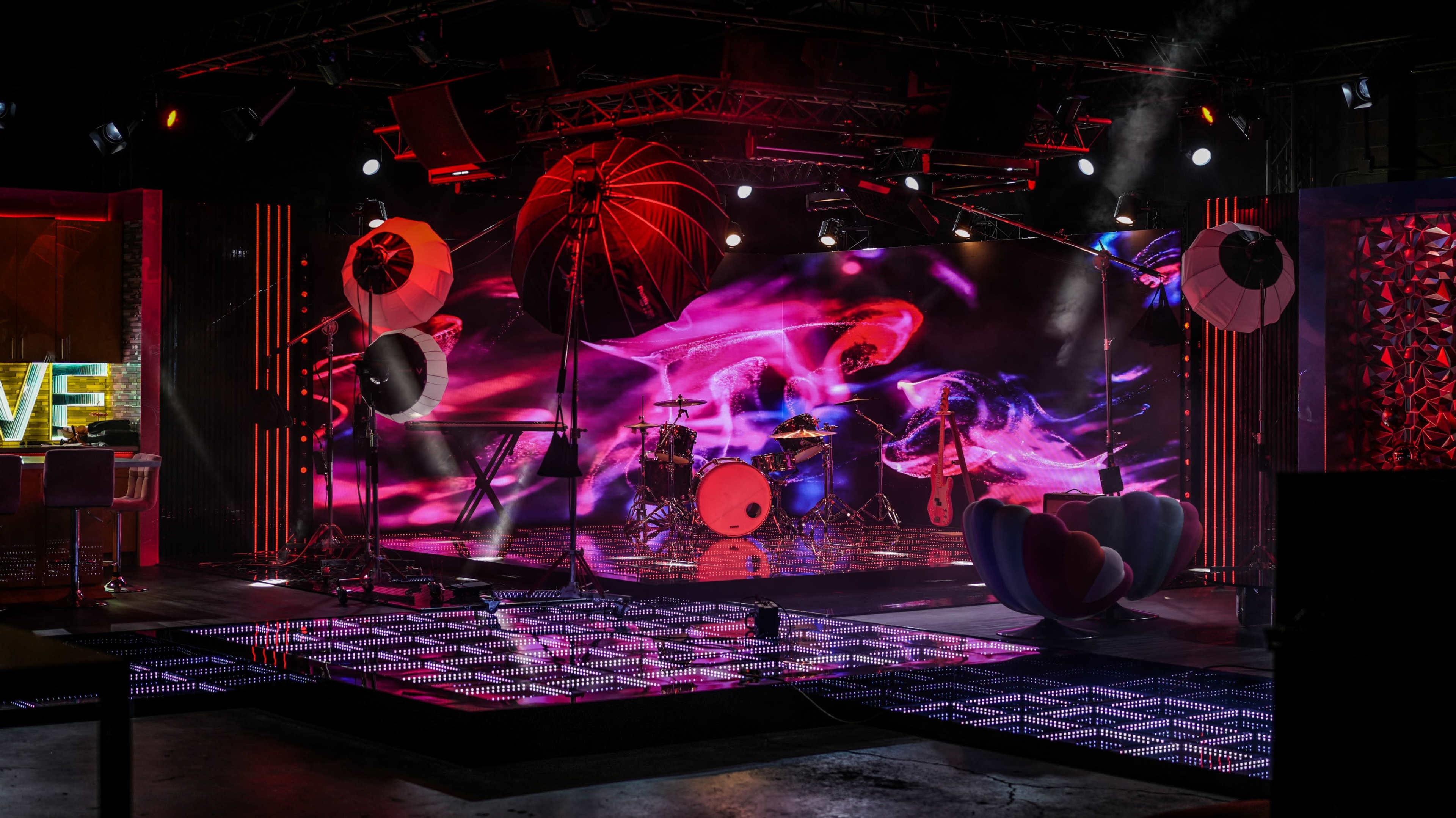 The image shows a vibrant stage setup with colorful lighting, musical instruments, and a large multimedia backdrop, ready for a live performance.