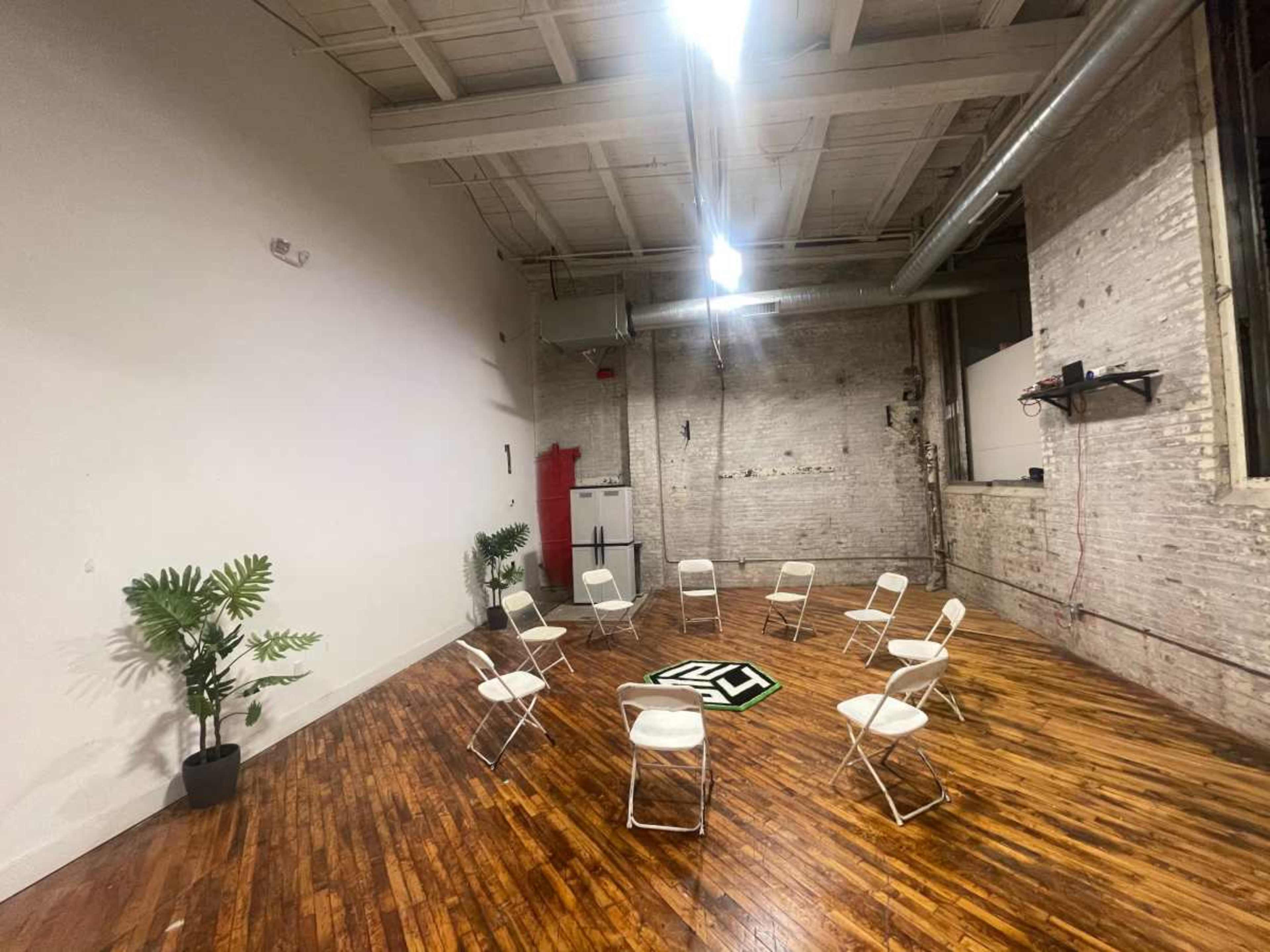 The image shows a spacious room with wooden flooring, featuring a circle of white folding chairs around a black and white patterned mat, with an artificial plant in a pot.