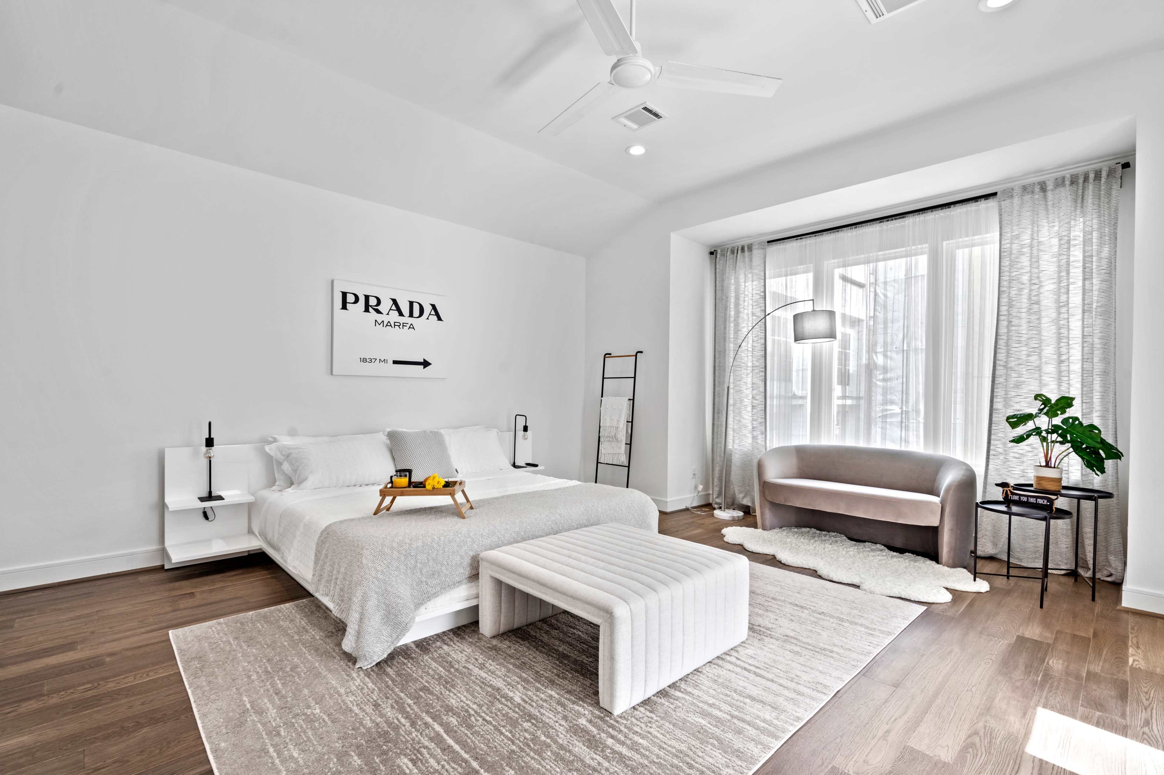 A minimalist bedroom featuring a white bed, a hanging sign with "PRADA," a modern armchair, a plant, and large windows with sheer curtains.