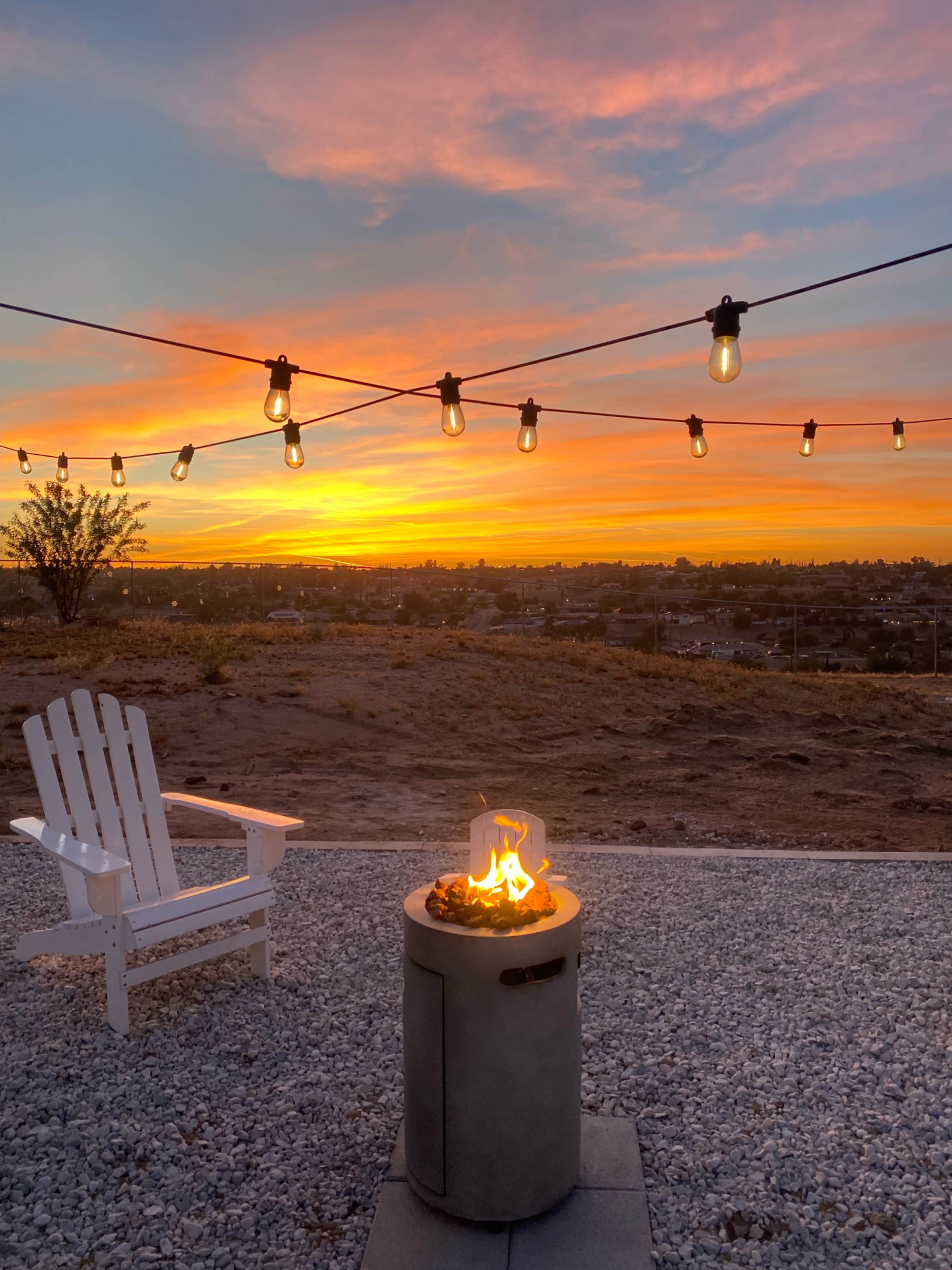 A fire pit surrounded by stones sits in front of an Adirondack chair under string lights, with a sunset illuminating the sky in the background.