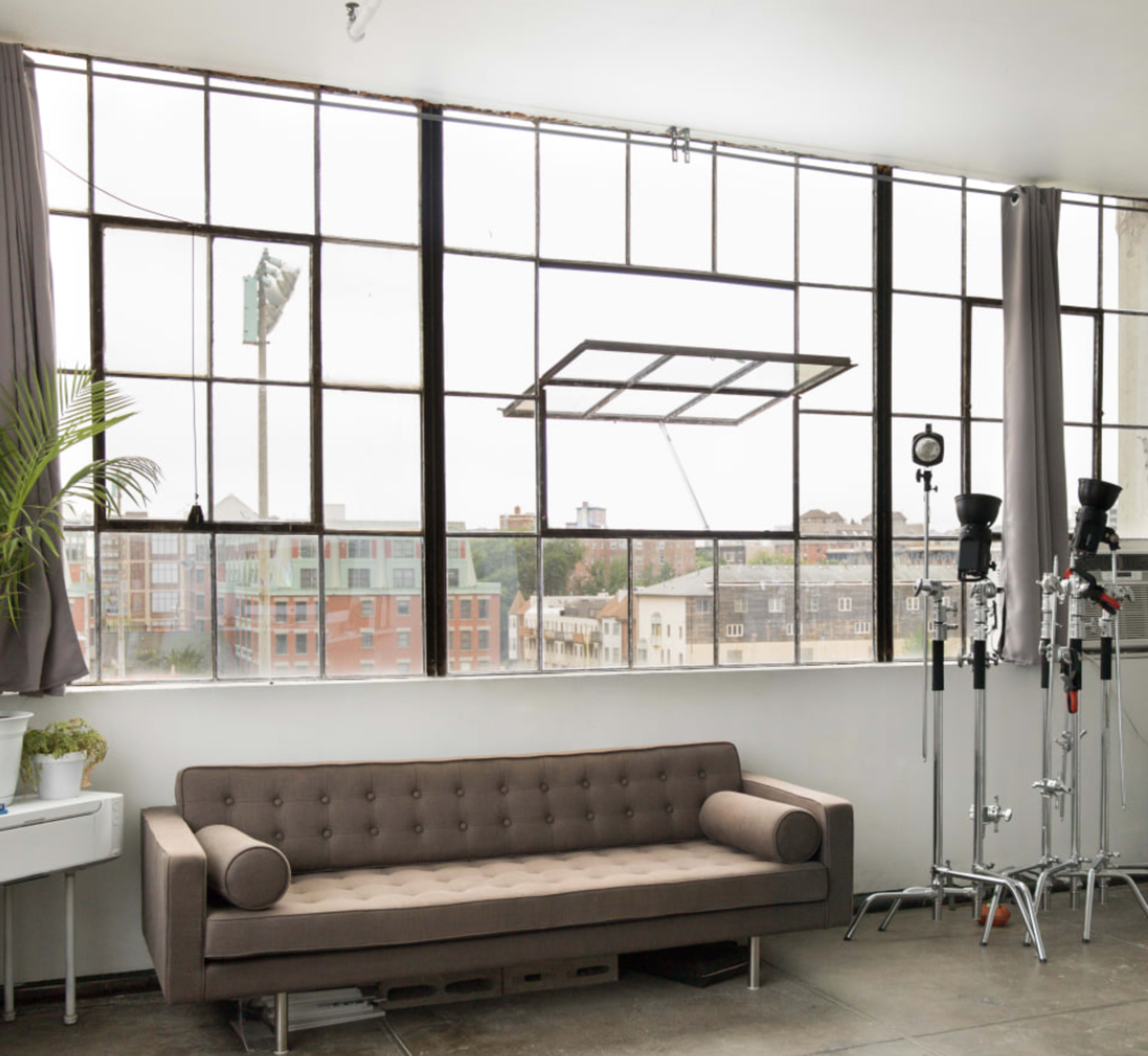 The image shows a spacious room with large windows, a brown couch, and studio equipment located near the window.