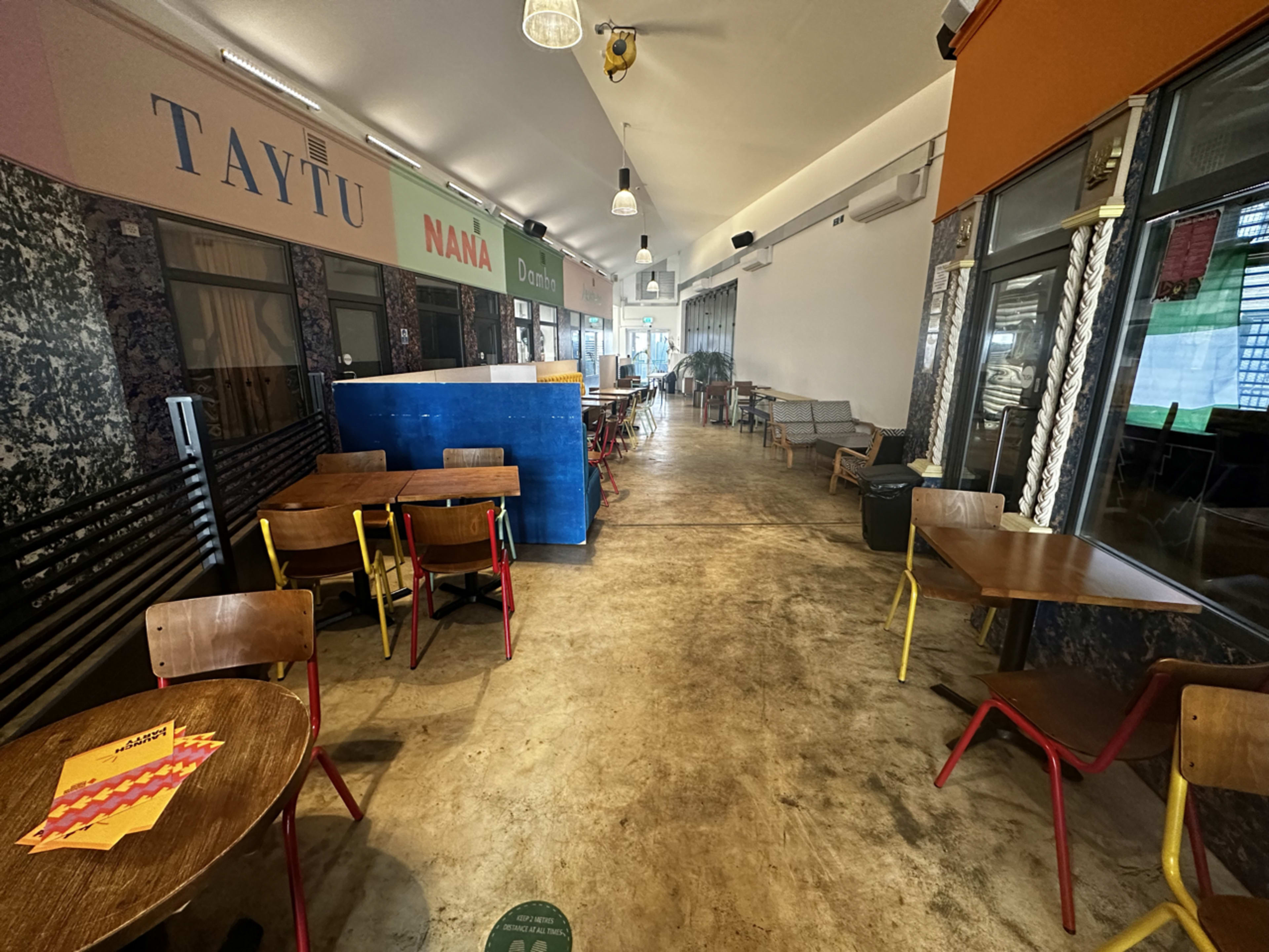 The image shows a mostly empty corridor of a dining area with a row of small wooden tables and chairs, featuring colorful walls and a blue counter at the end.