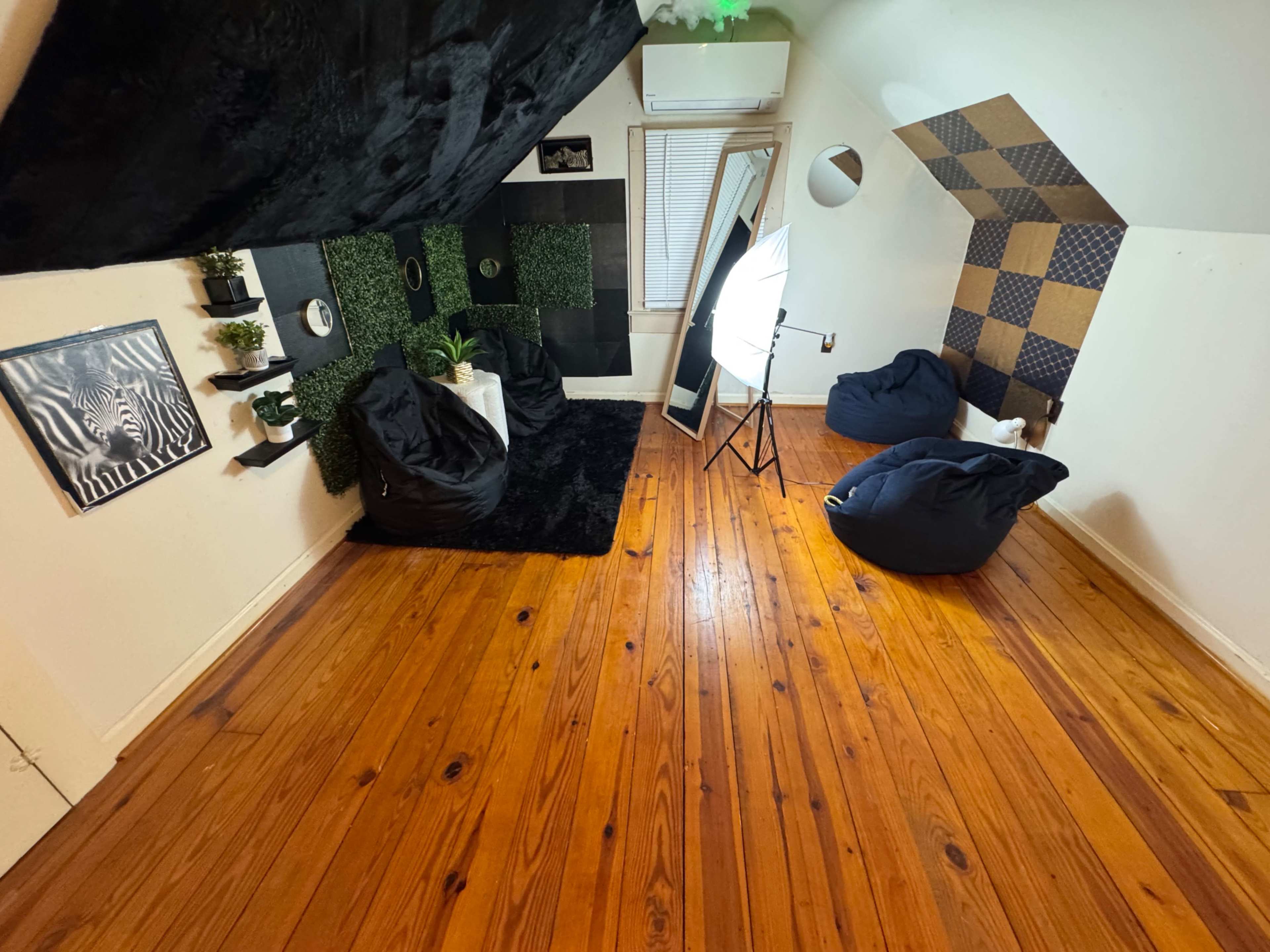 The image shows a cozy attic room with wooden floors, bean bags, a black rug, and a green wall display, along with a mirror and a softbox light set up for photography.