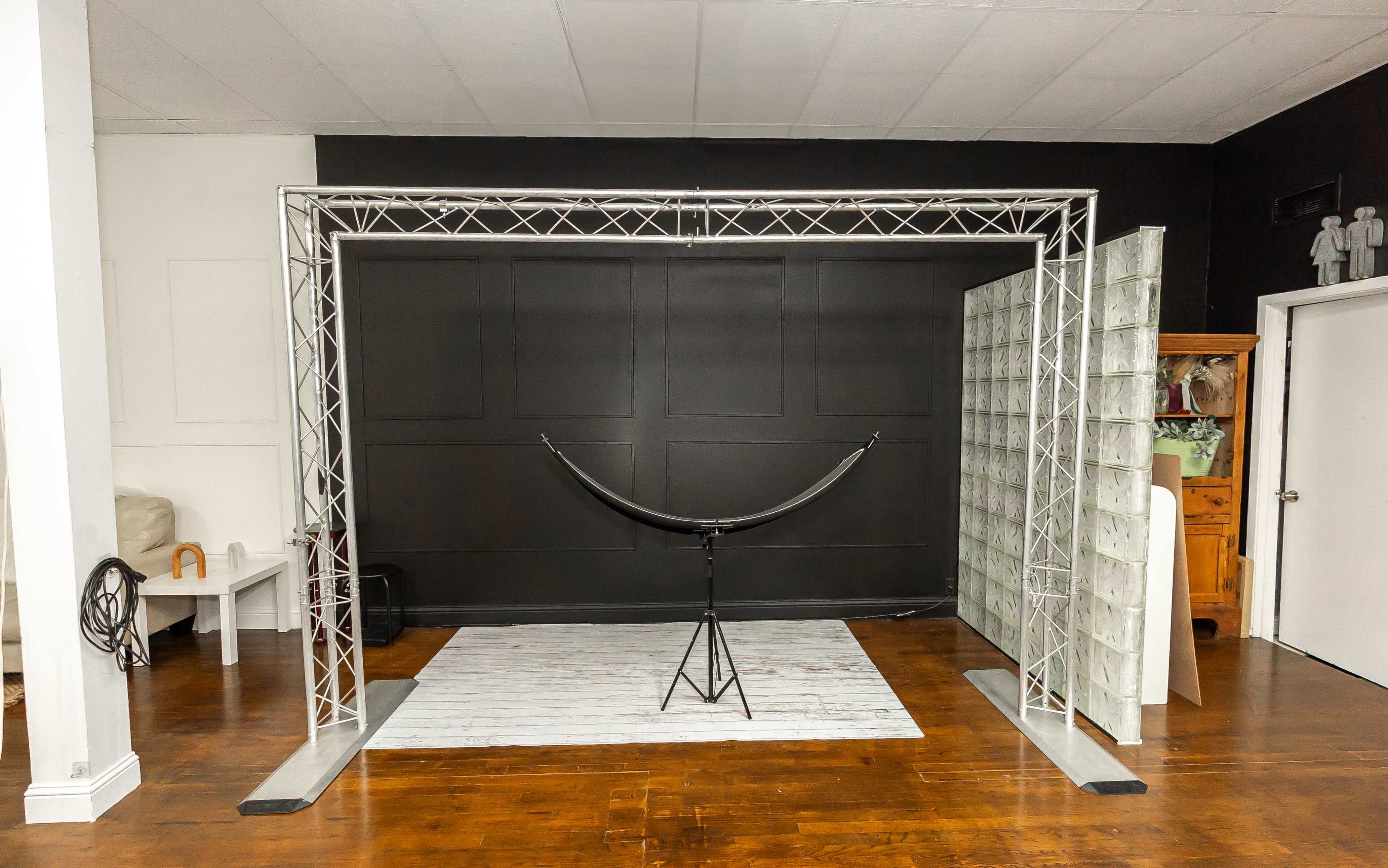 The image shows a photography setup with a metal truss frame, a curved reflector, and a white backdrop on a wooden floor in a minimally decorated studio space.