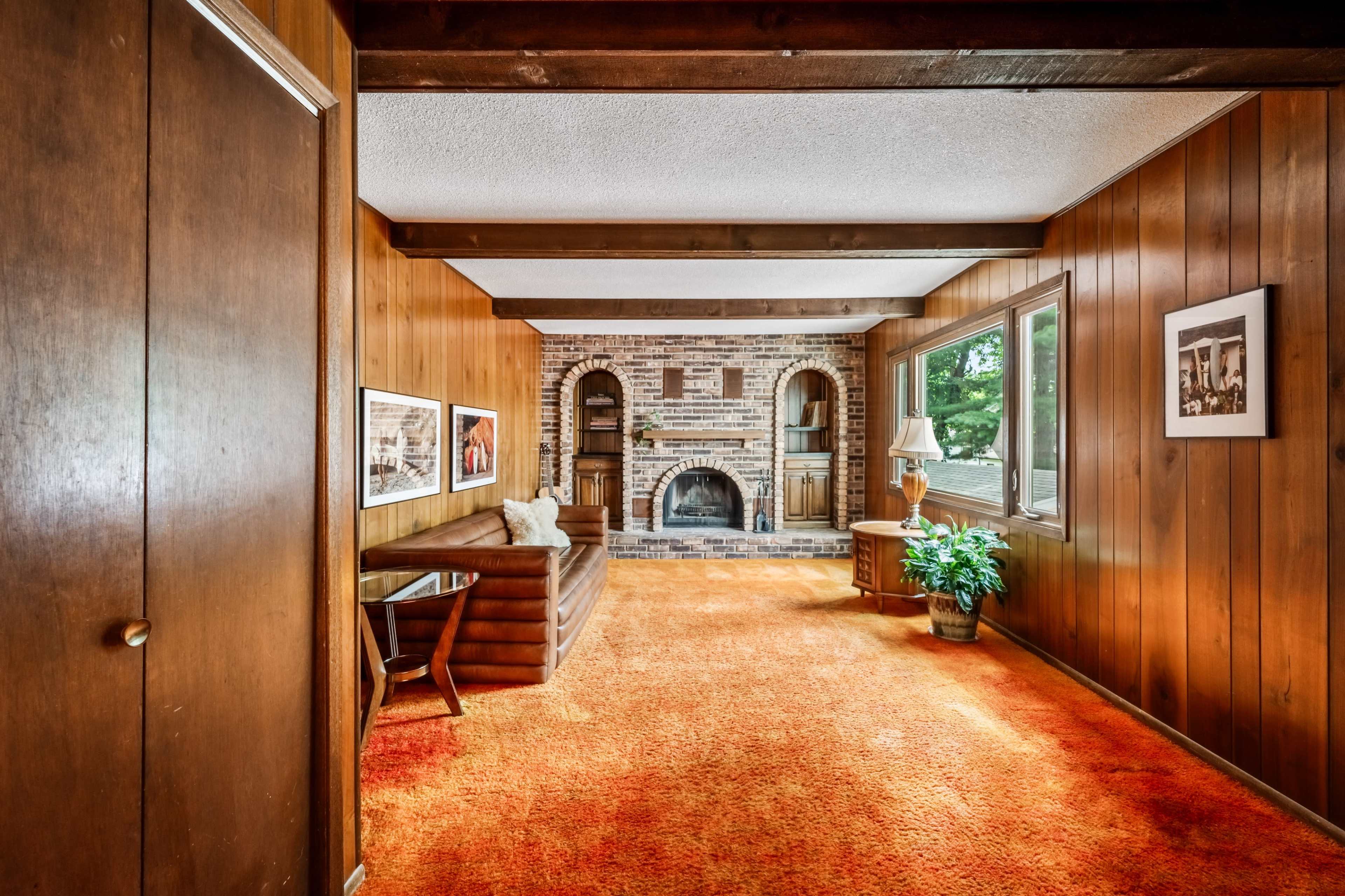 A living room with wood-paneled walls, a brick fireplace, and orange carpeting.