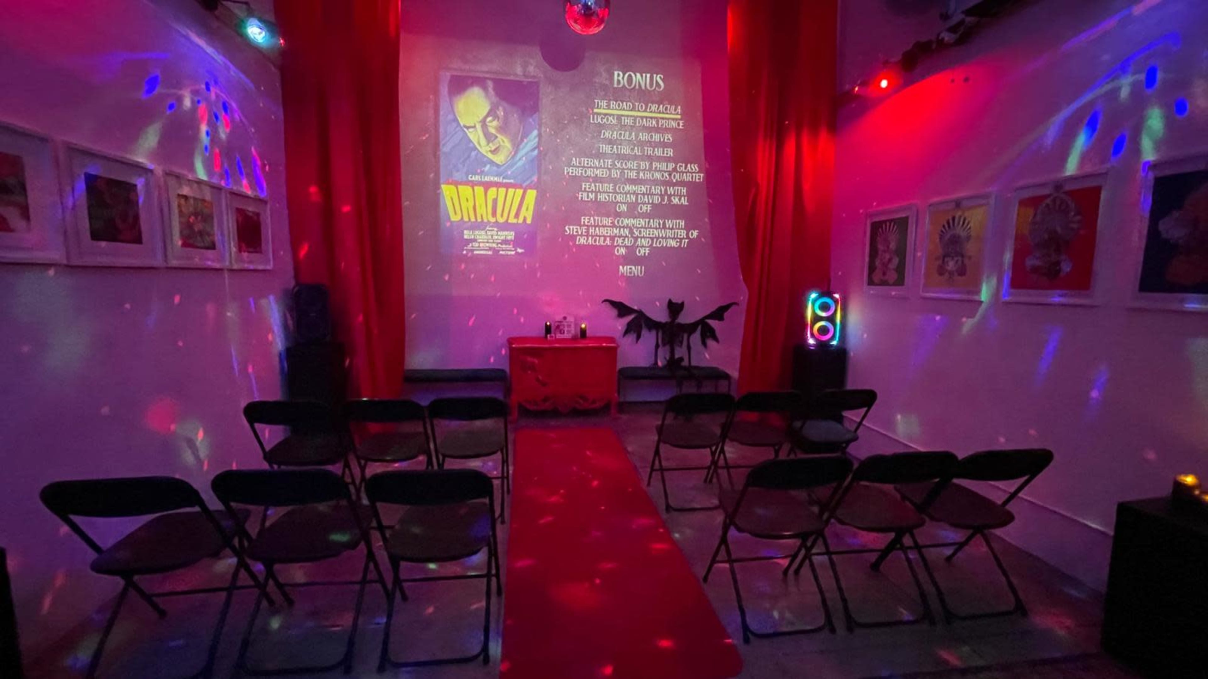The image shows a dimly lit room set up for an event, featuring rows of chairs facing a stage with a red curtain, a table adorned with a Dracula-themed display, and colorful lights projecting patterns on the walls.