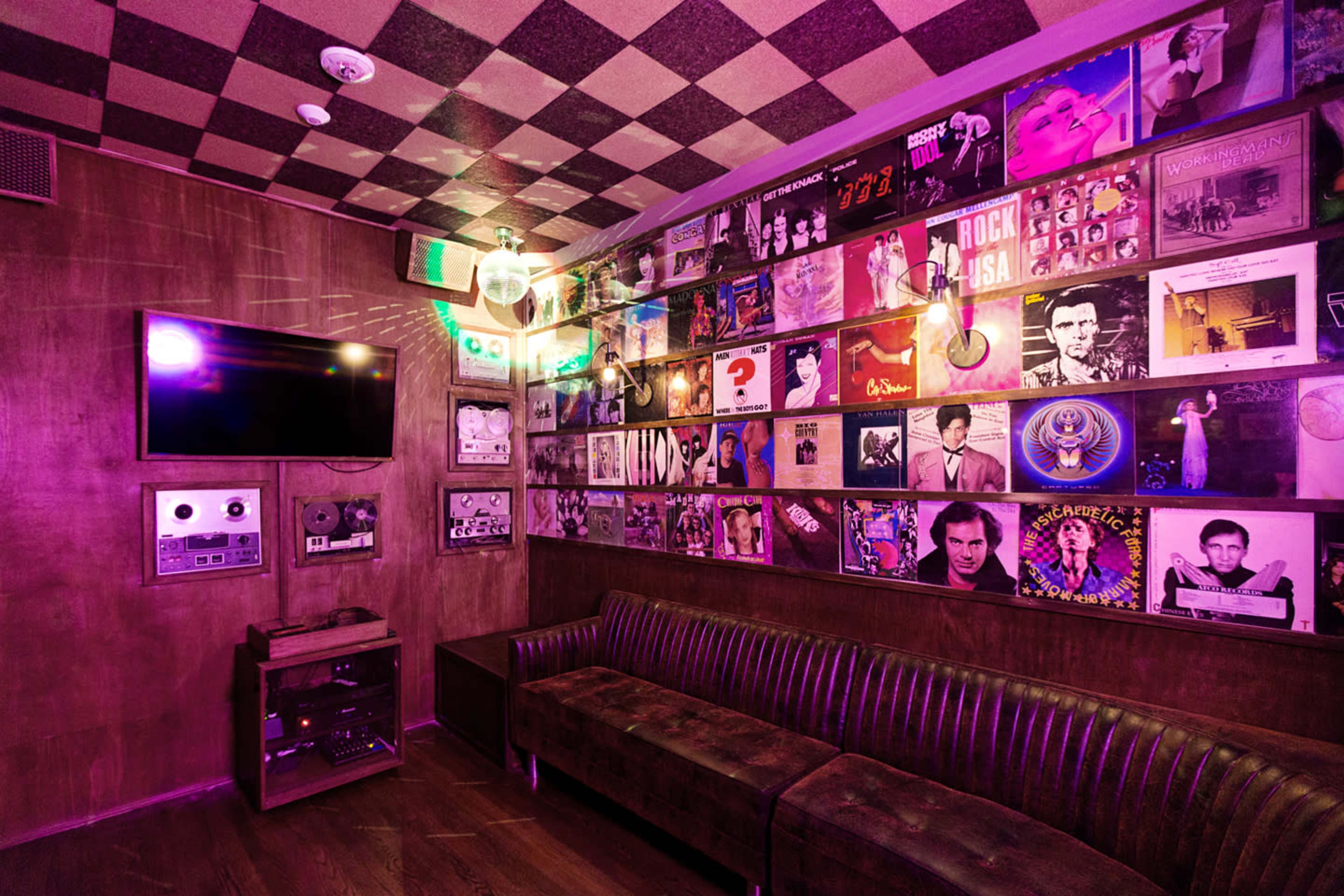 The image shows a small, dimly lit room with a couch, decorated with a wall of framed vinyl records and a television mounted on the wall.