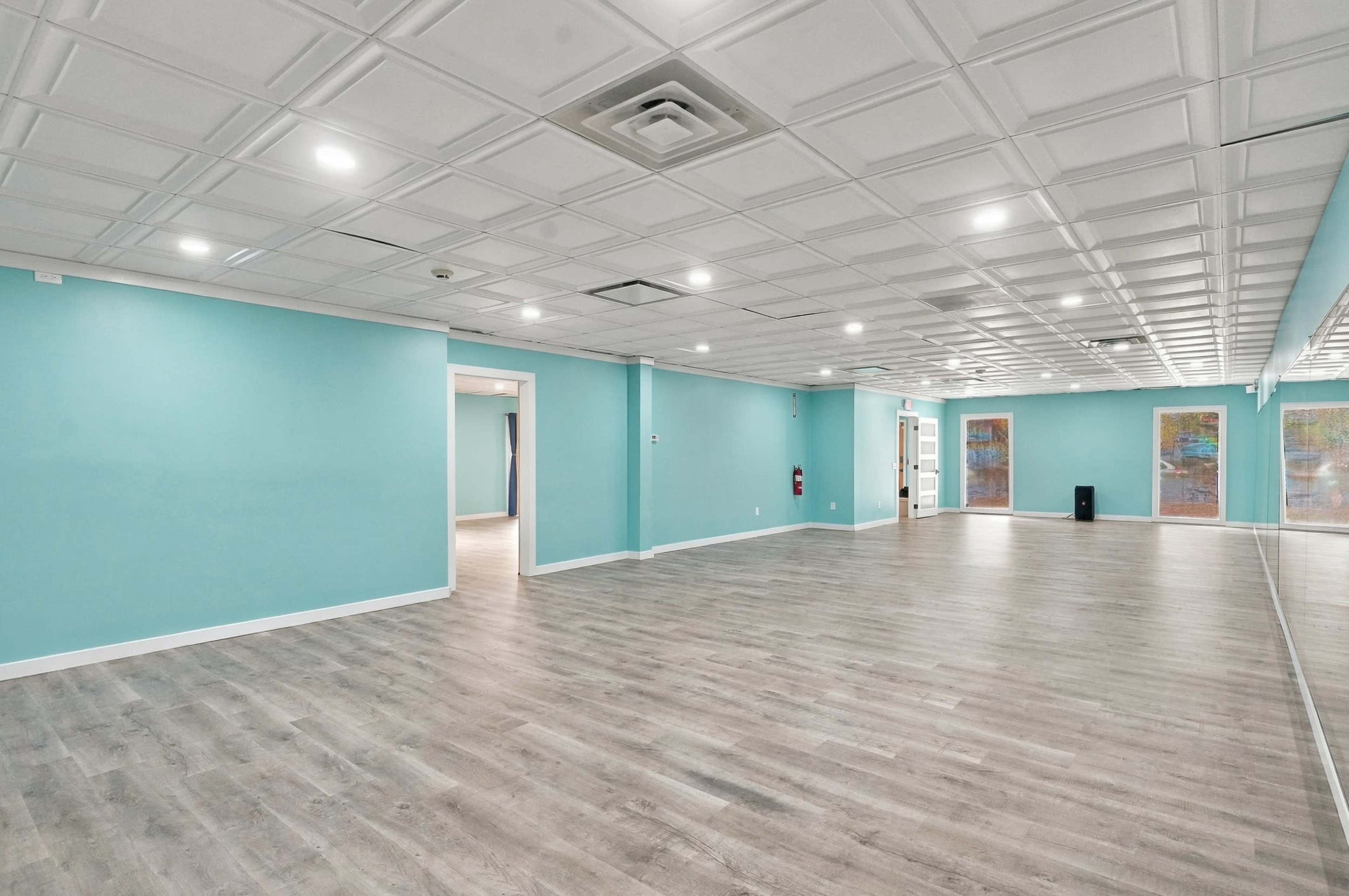 The image shows a spacious, empty room with light blue walls, a hardwood floor, and overhead lighting, featuring a mirrored section on one side.